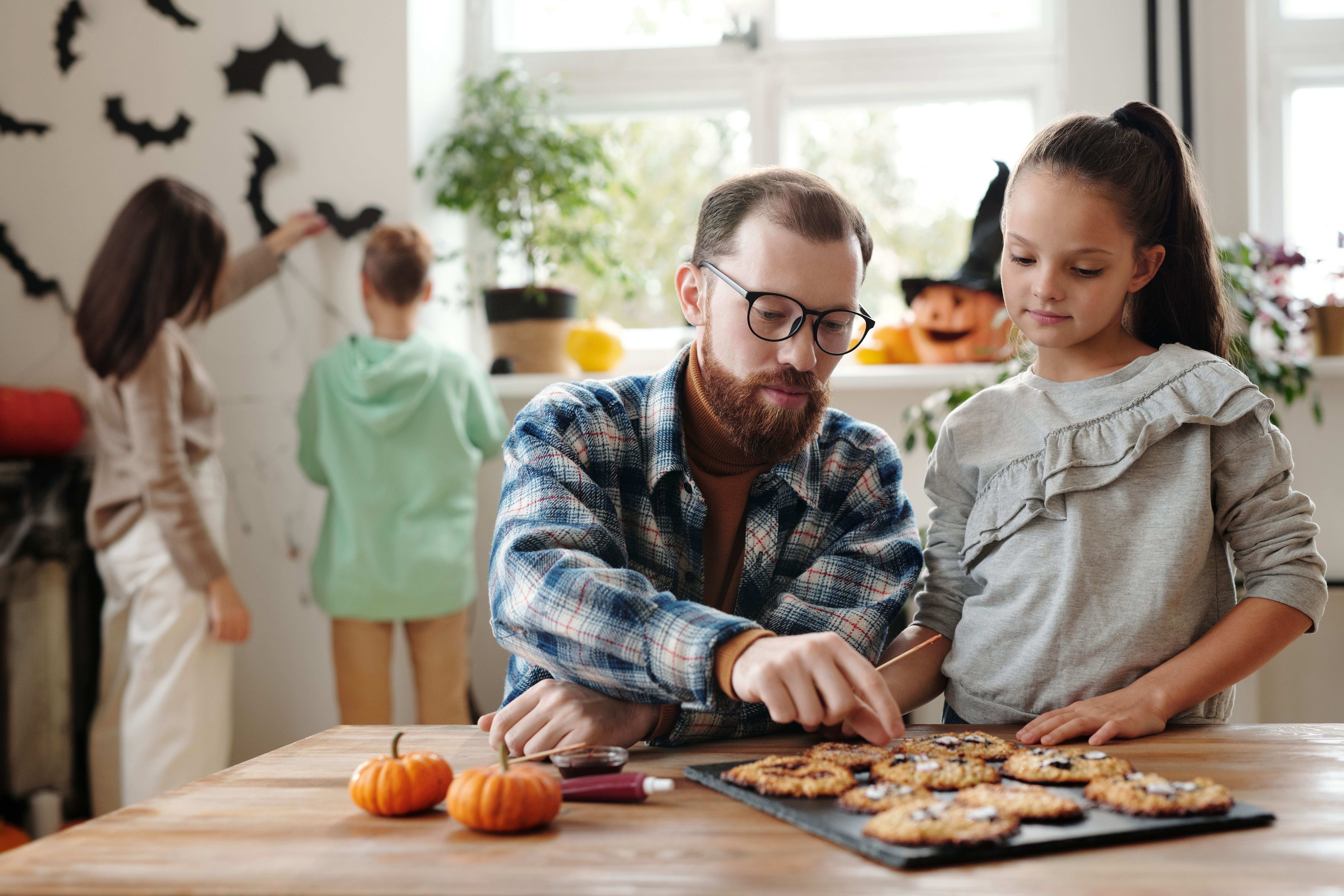 A father and daughter decorating Halloween cookies together at a kitchen table while family members decorate the wall in the background.