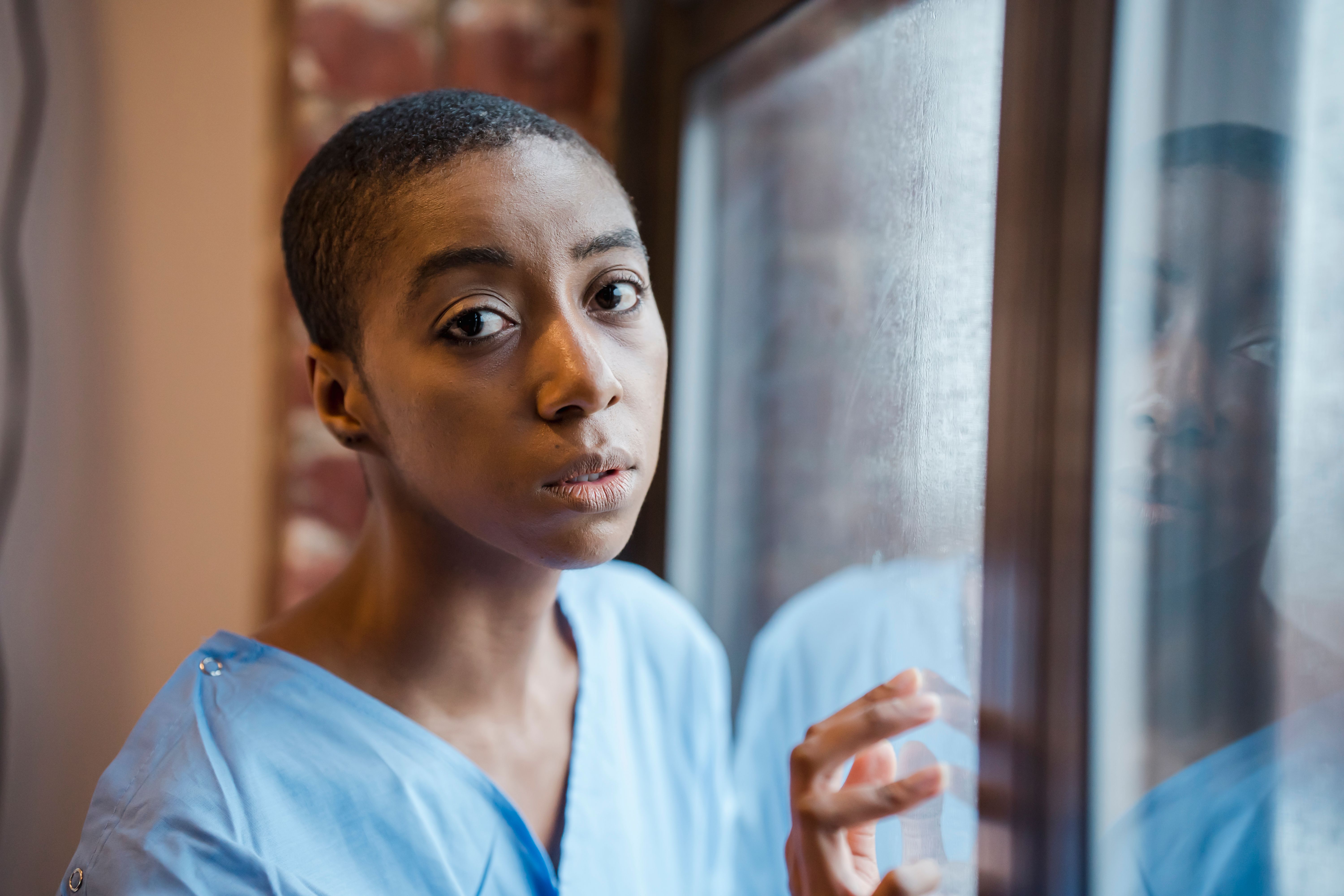 A woman in a light blue medical gown touching a glass window while looking thoughtfully into the camera.