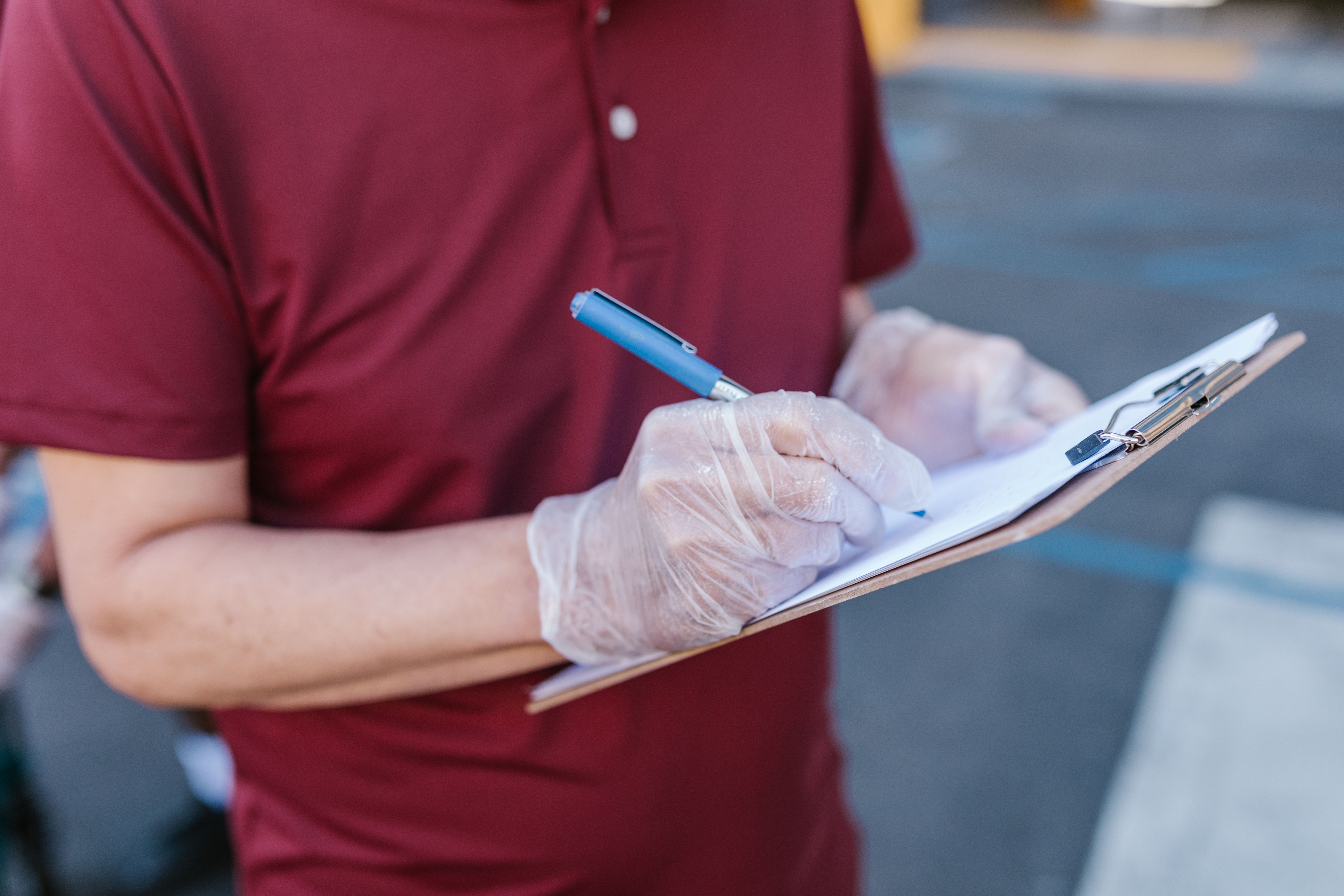 A person in a maroon shirt wearing disposable gloves writing on a clipboard with a pen.