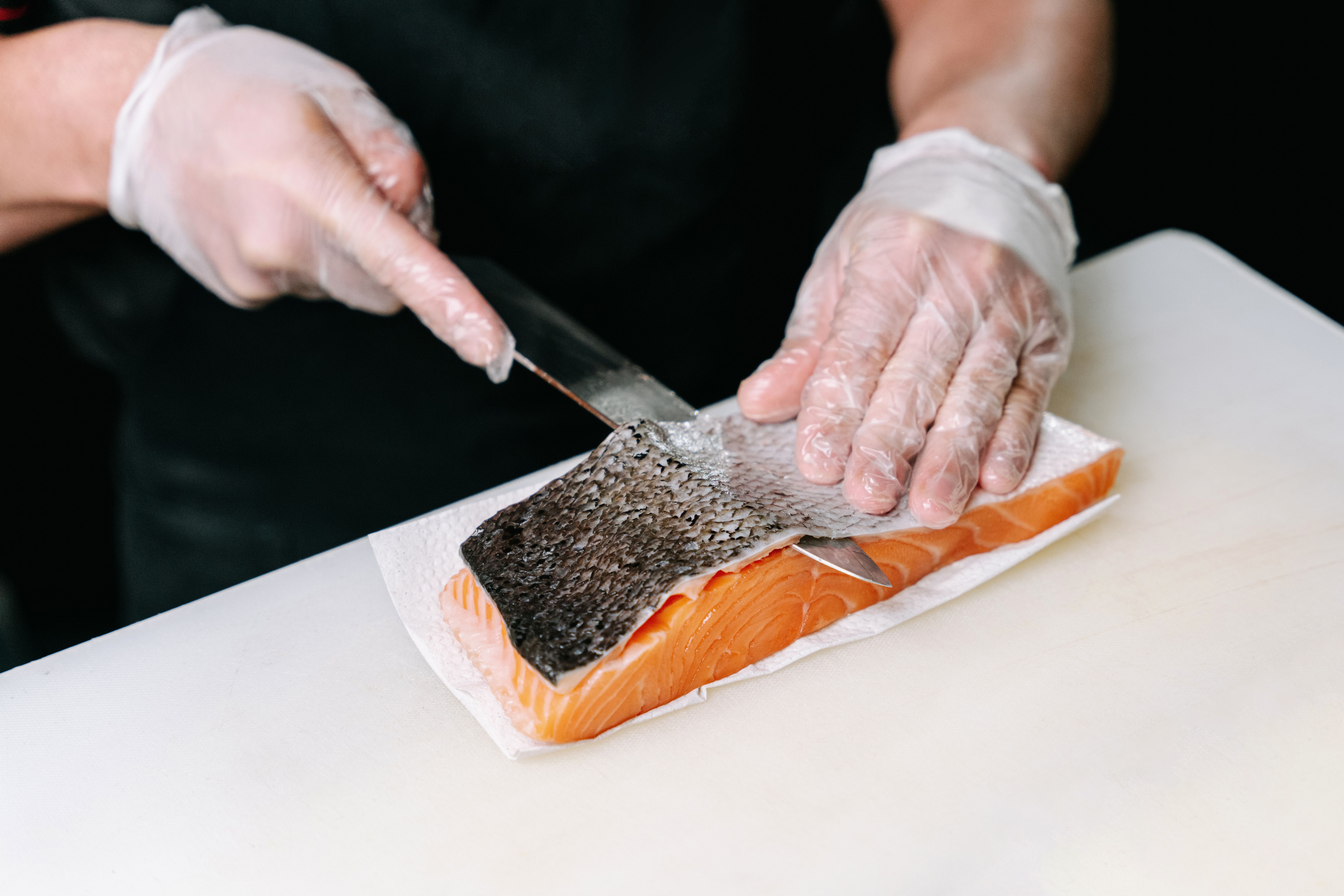 Close-up of a chef wearing clear plastic gloves using a sharp knife to carefully skin a salmon fillet on a white cutting board.