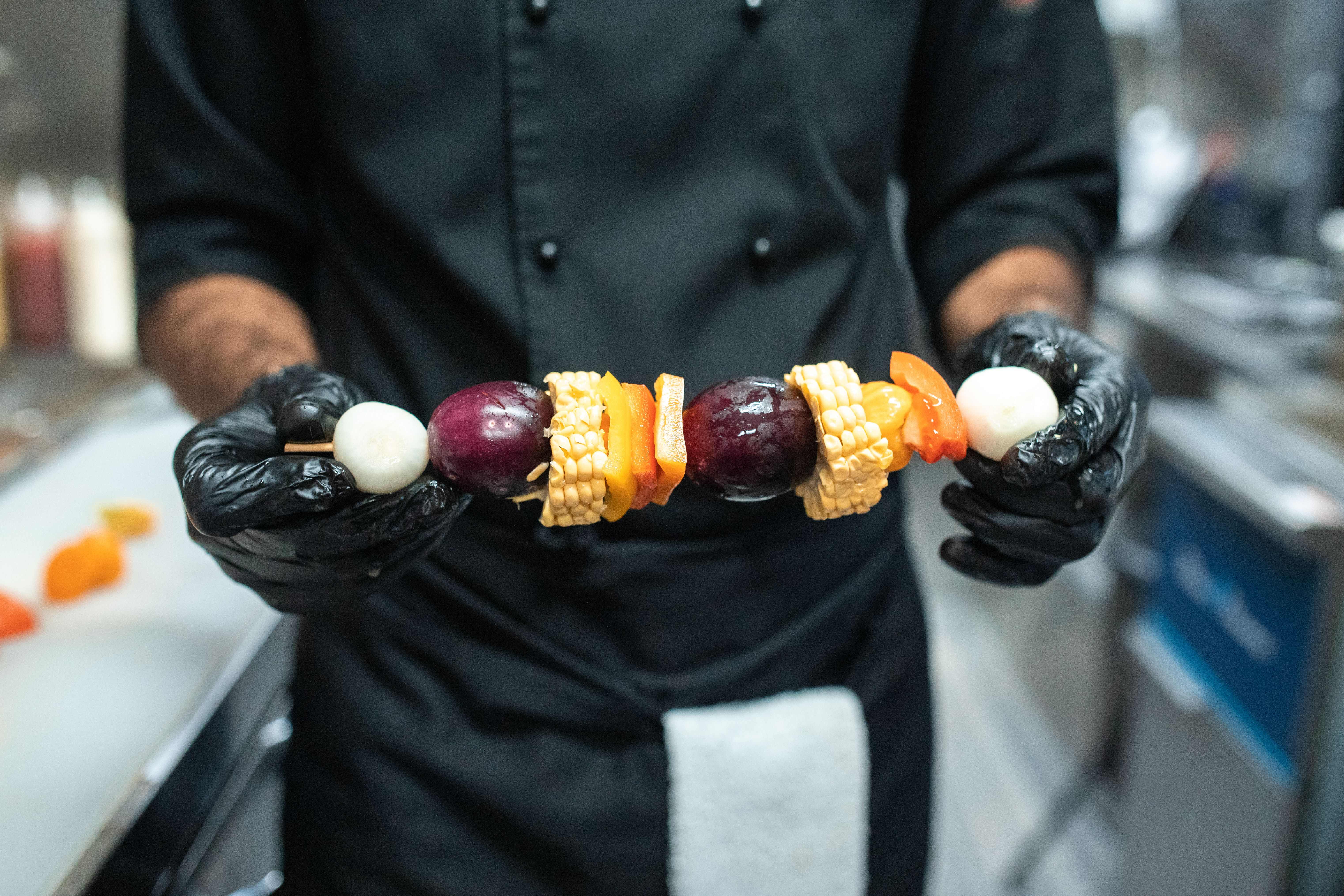 Close-up of a chef wearing black disposable gloves holding a wooden skewer stacked with colorful vegetables and onions.
