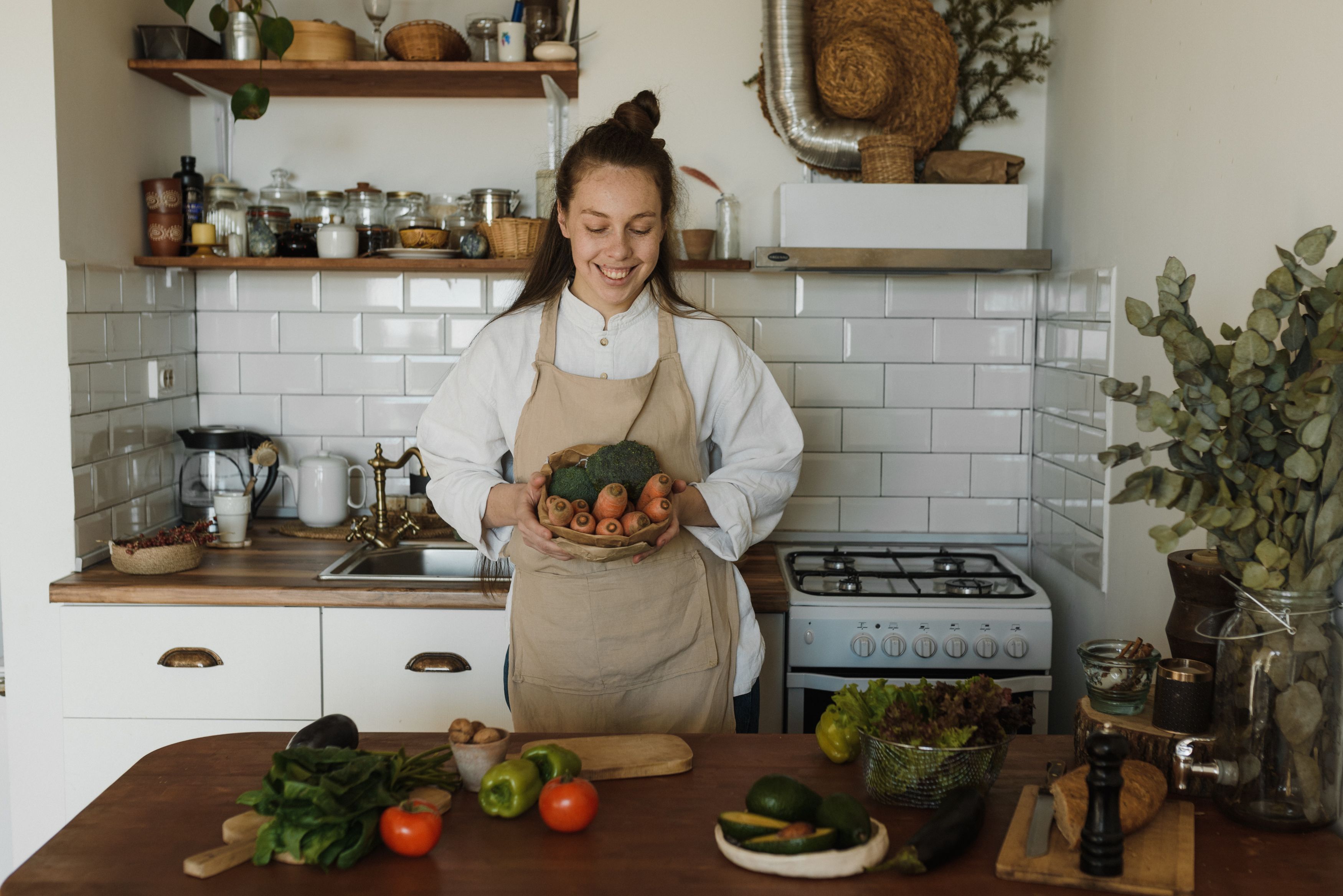 A smiling woman wearing a beige apron standing in a cozy kitchen holding a paper bowl filled with fresh broccoli and carrots.