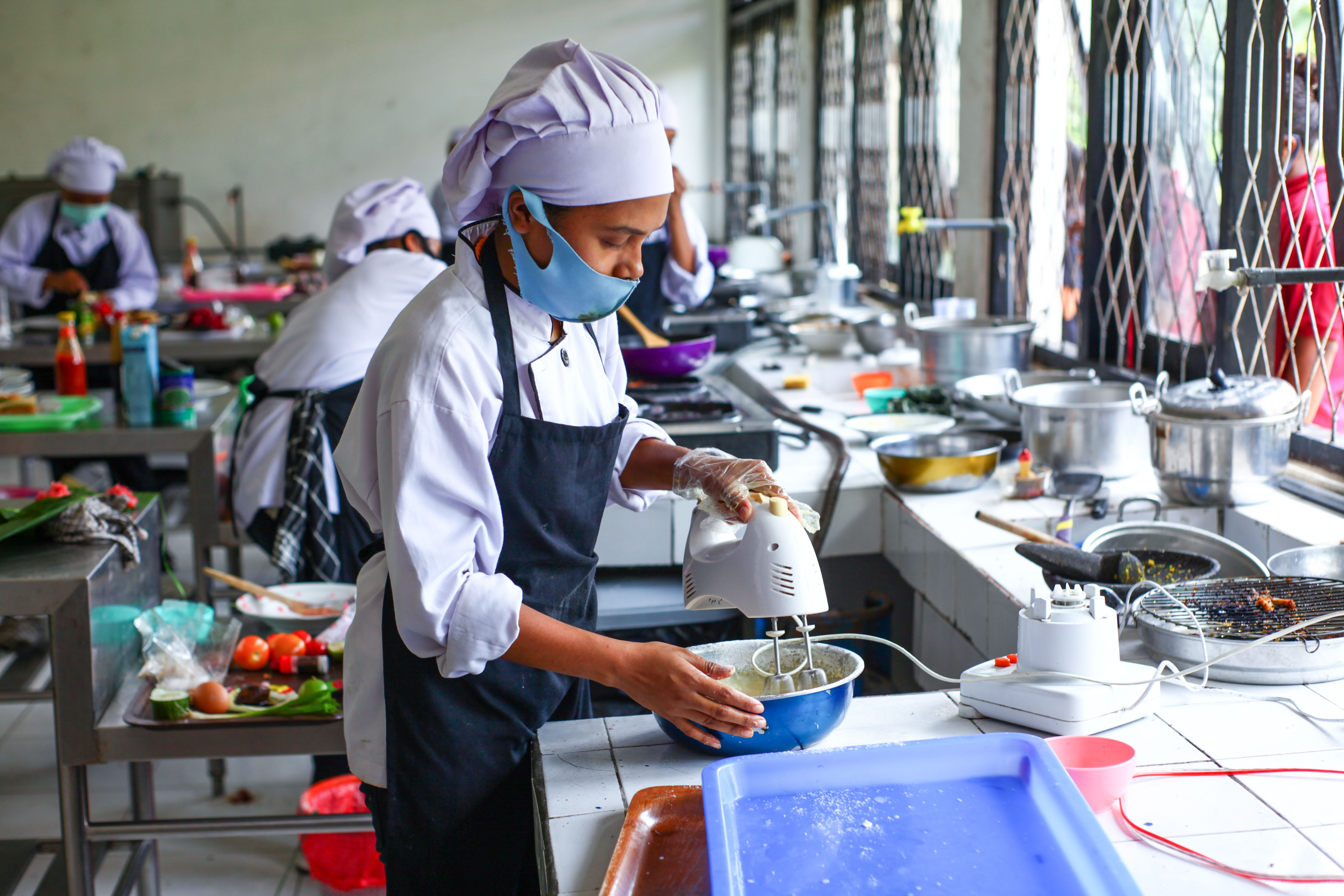 Trainee chefs in white chef hats and face masks working at kitchen counters mixing batter with an electric hand mixer.