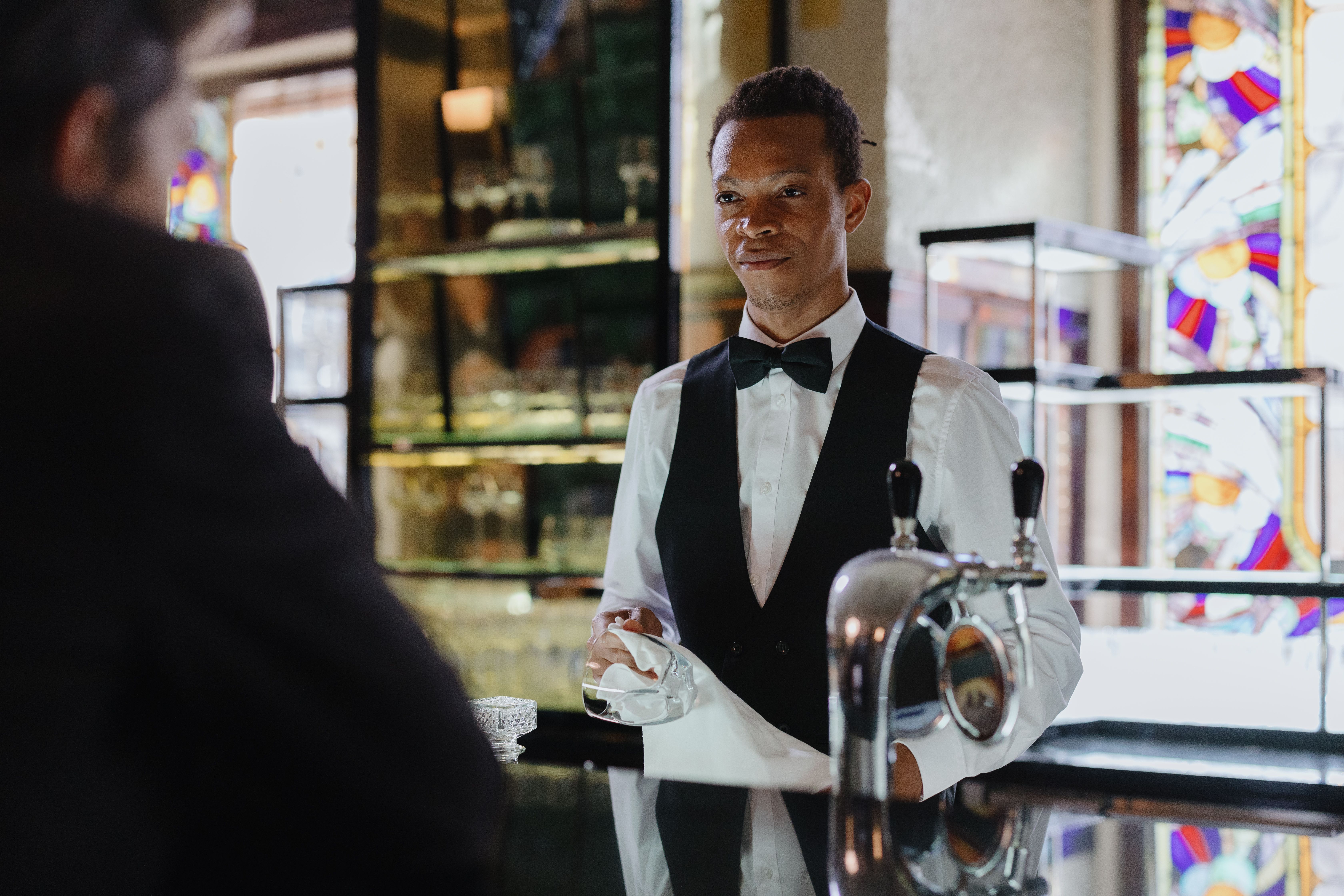 A bartender wearing a vest and bow tie polishing a glass behind a bar counter with beer taps.