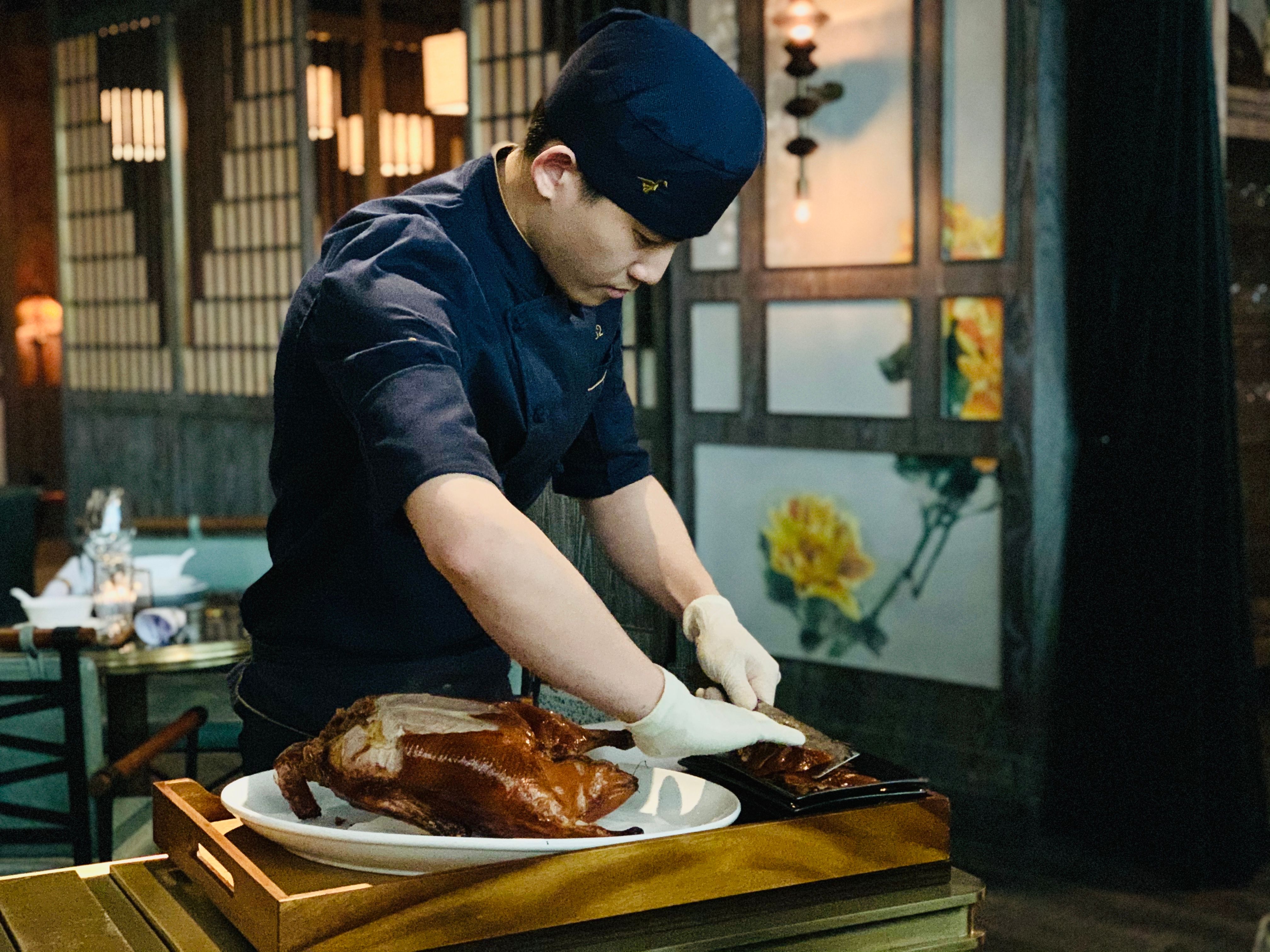 A chef wearing a blue cap and white gloves carving a roasted duck at a dining table.