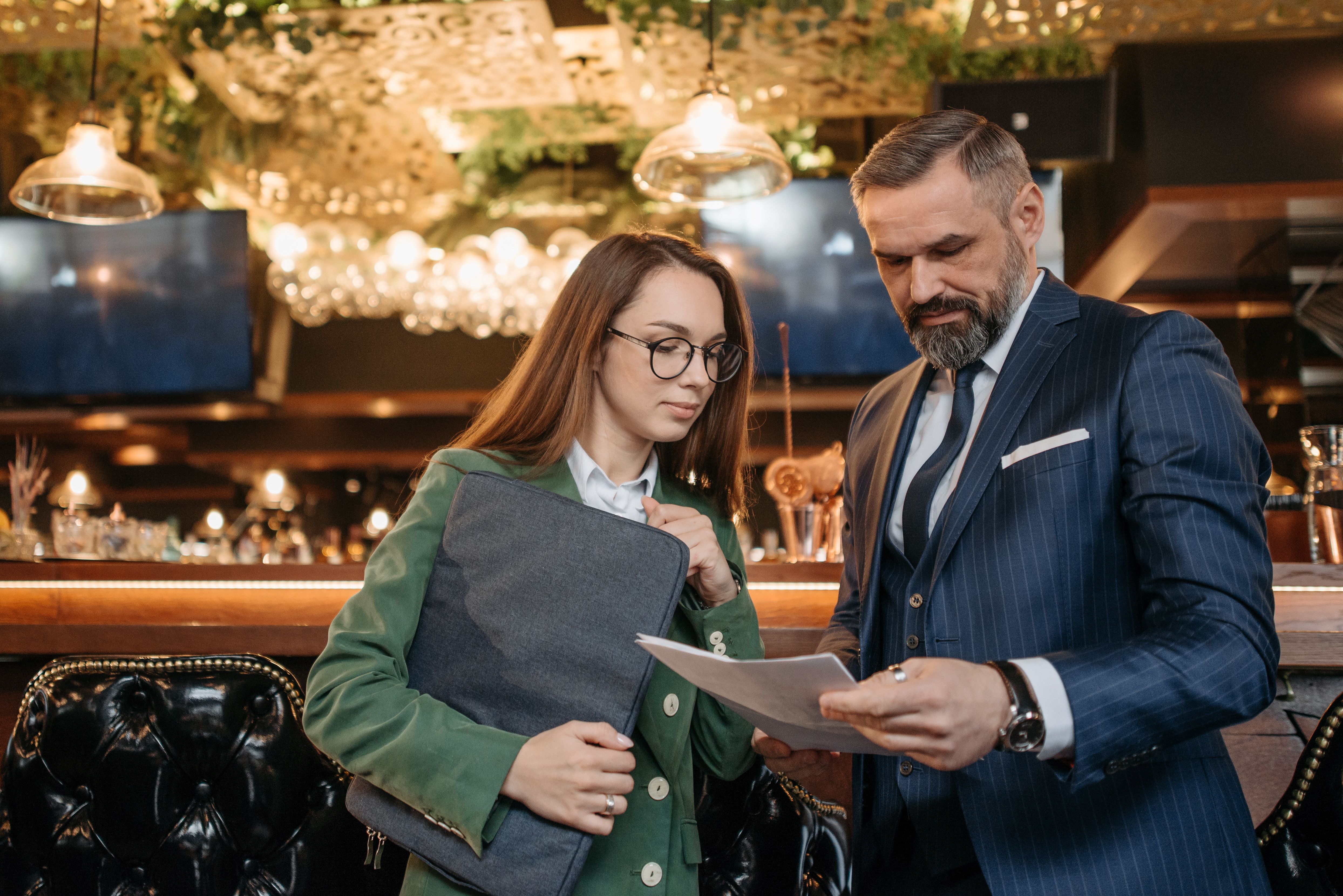 A woman in glasses holding a folder and a man in a navy suit reviewing printed documents inside a restaurant.