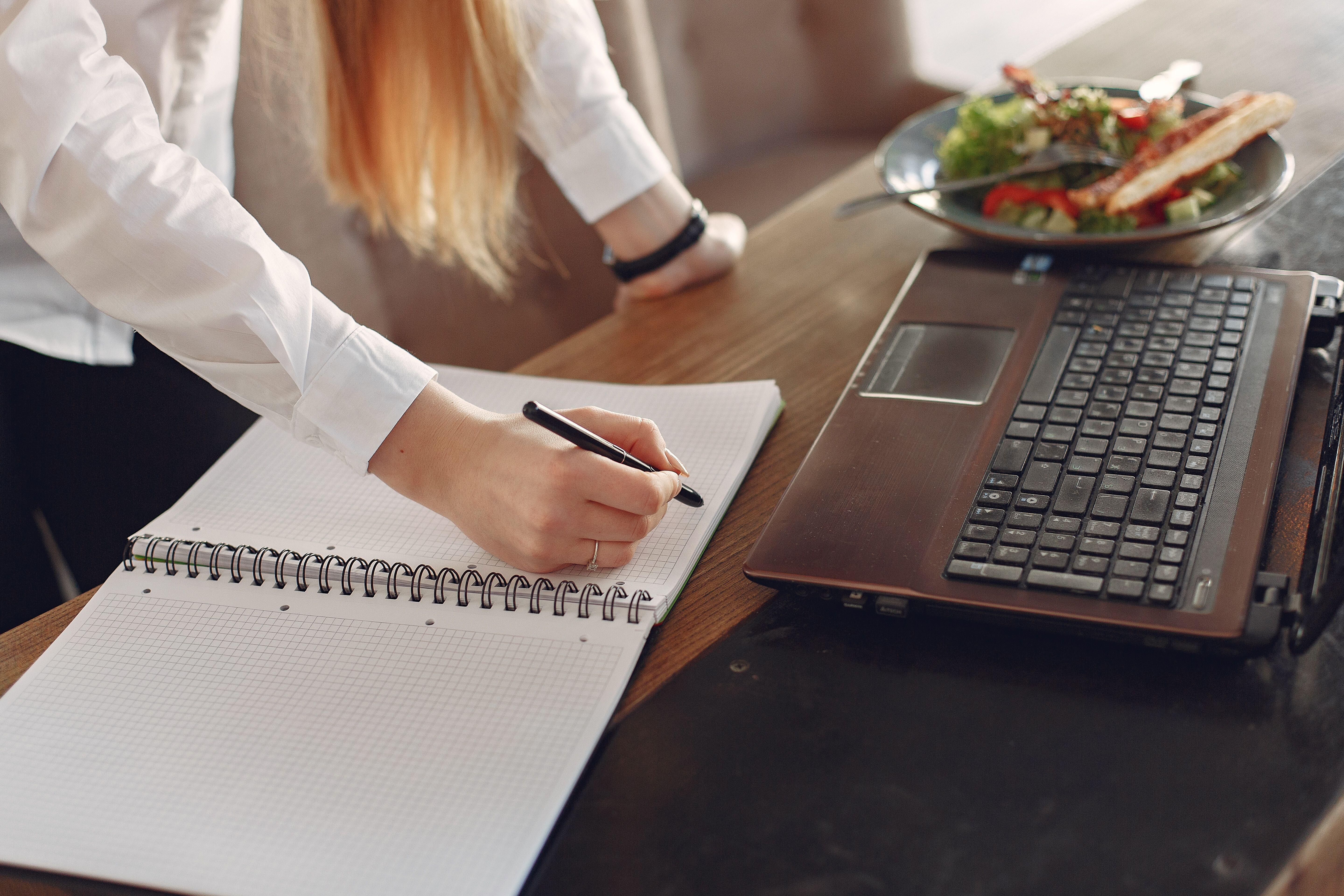 Close-up of a person writing in a spiral notebook with a pen next to an open laptop and a plate of salad.