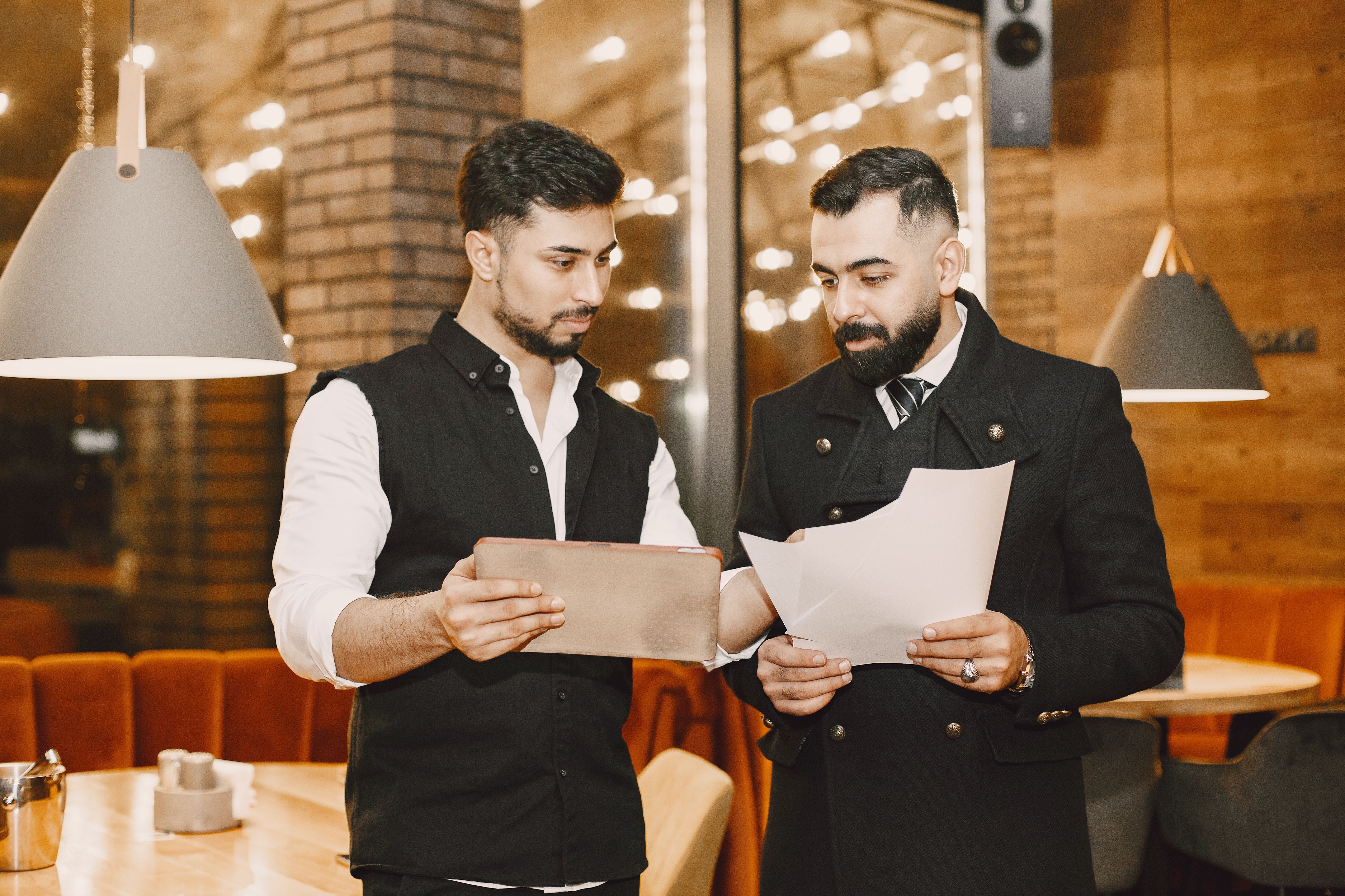 Two men standing in a restaurant looking at documents and a tablet together.