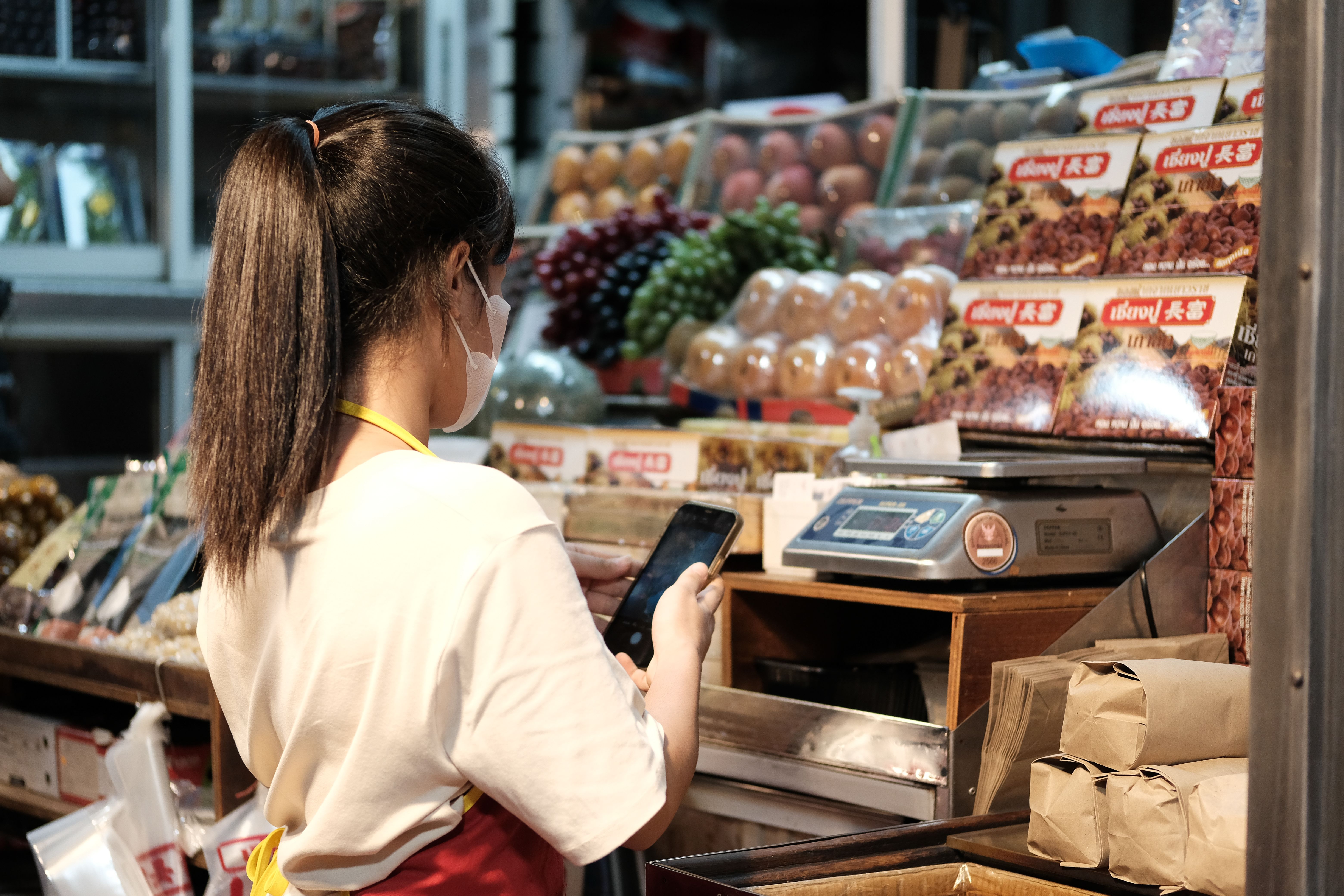 Side view of a market vendor in a face mask using a smartphone next to a digital scale and packaged goods.
