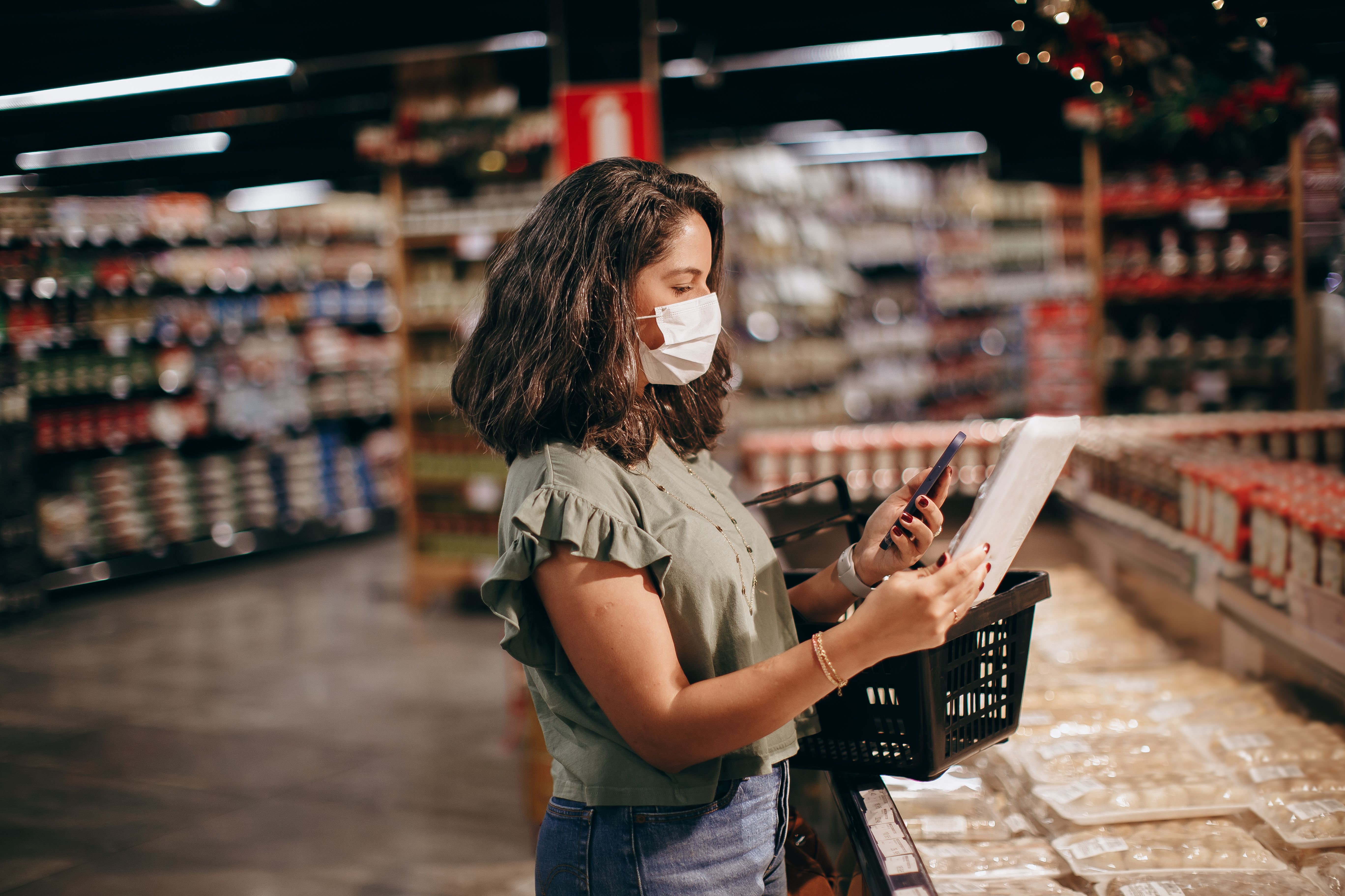A woman wearing a protective face mask holding a shopping basket and checking a food item using her smartphone in a grocery store aisle.