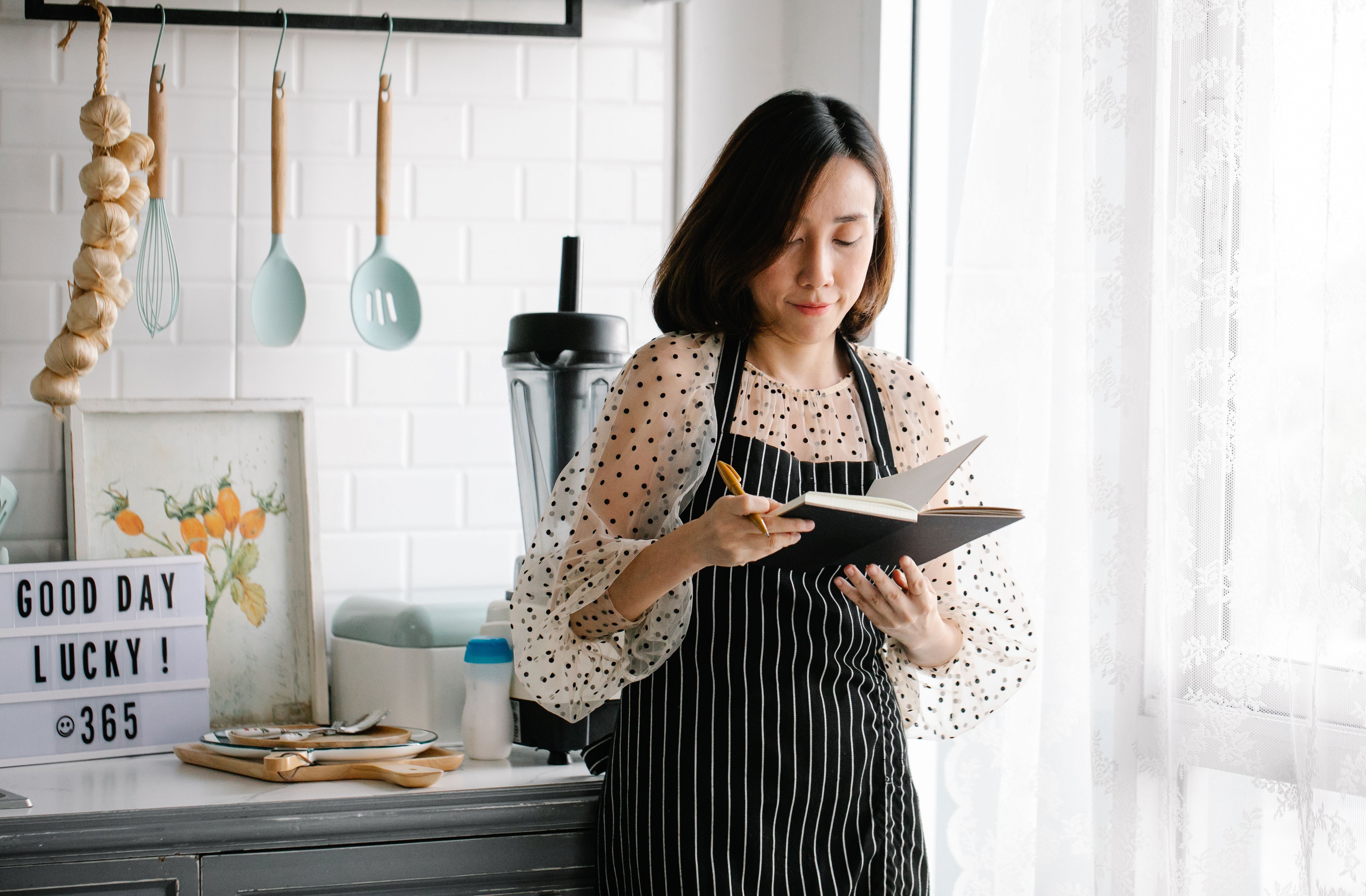 A woman wearing a striped apron standing in a kitchen reviewing notes in a small black notebook.