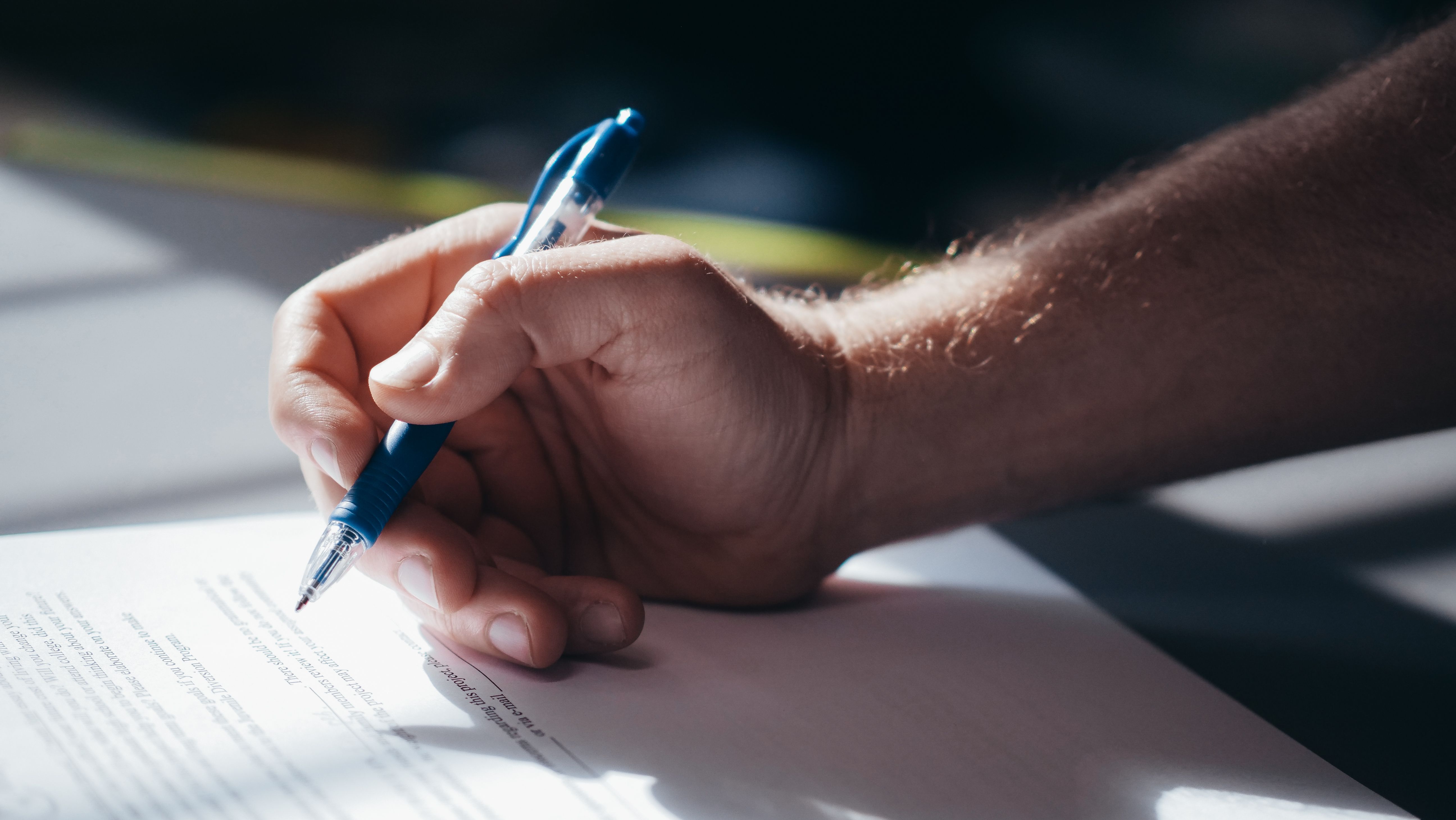 Close-up of a hand holding a blue pen signing or filling out a paper document on a desk.