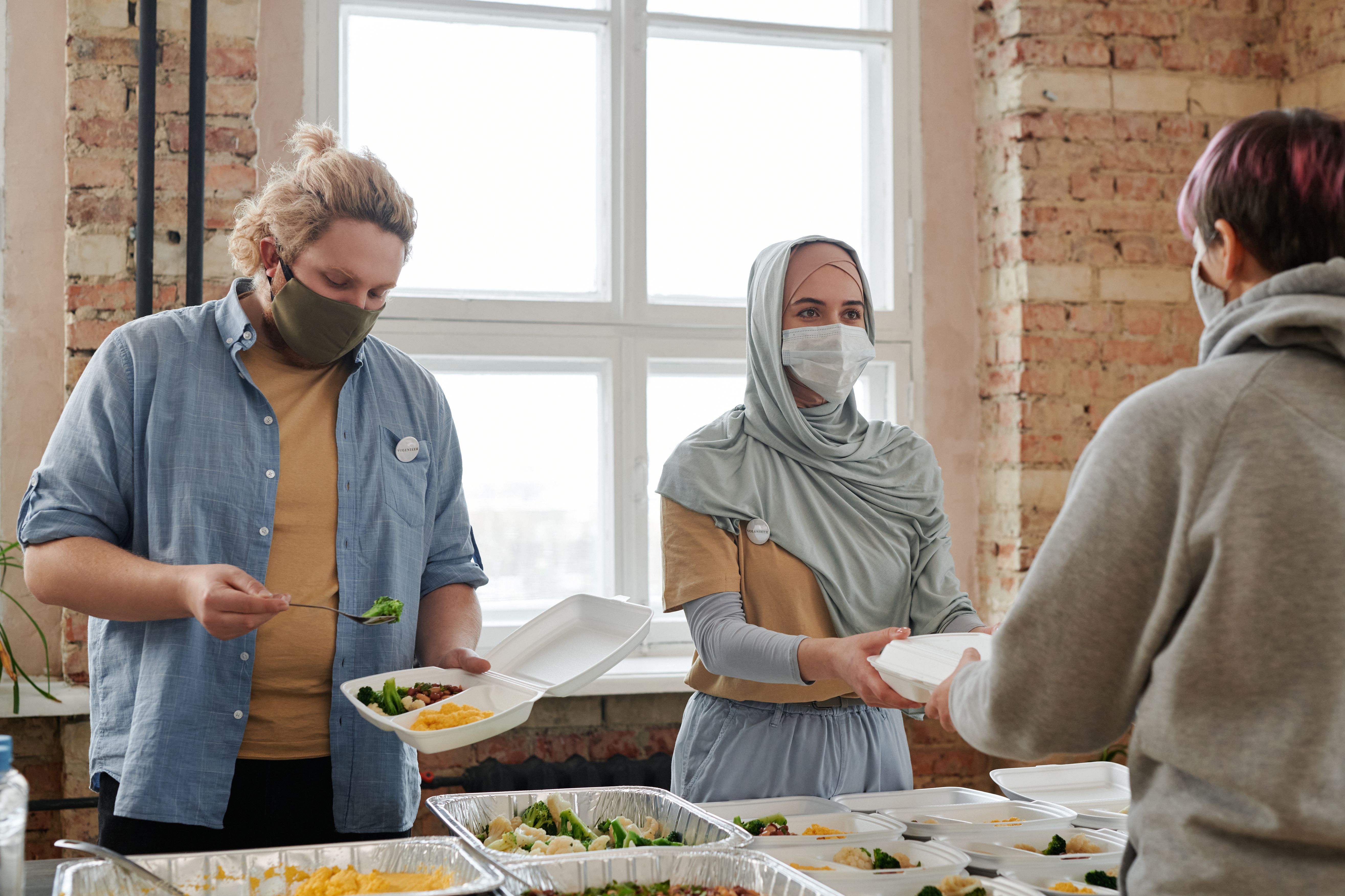 Volunteers wearing face masks packing and serving hot meals into takeaway containers from aluminum steam pans.