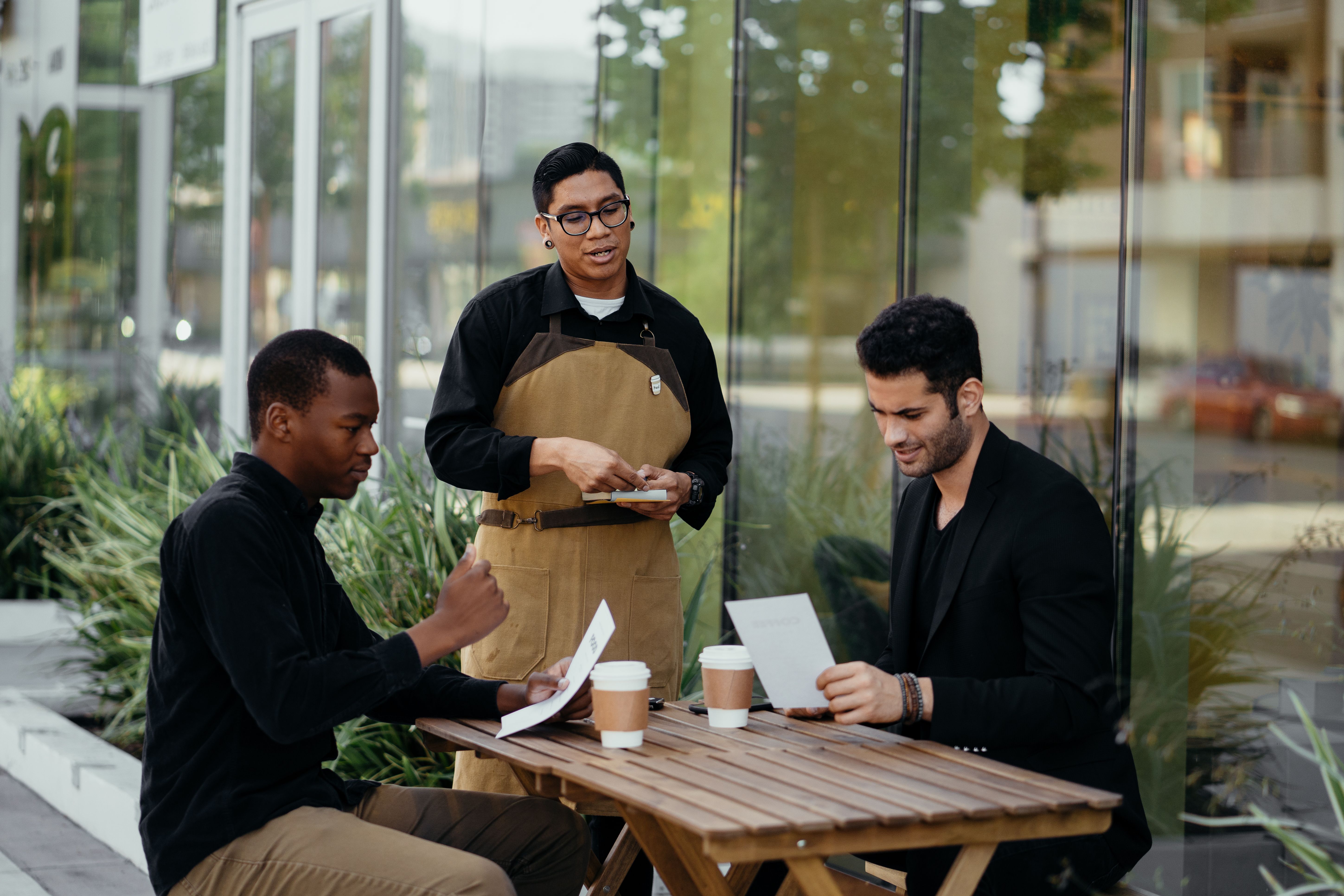 A waiter in a tan apron taking a order from two male customers seated at an outdoor café table.