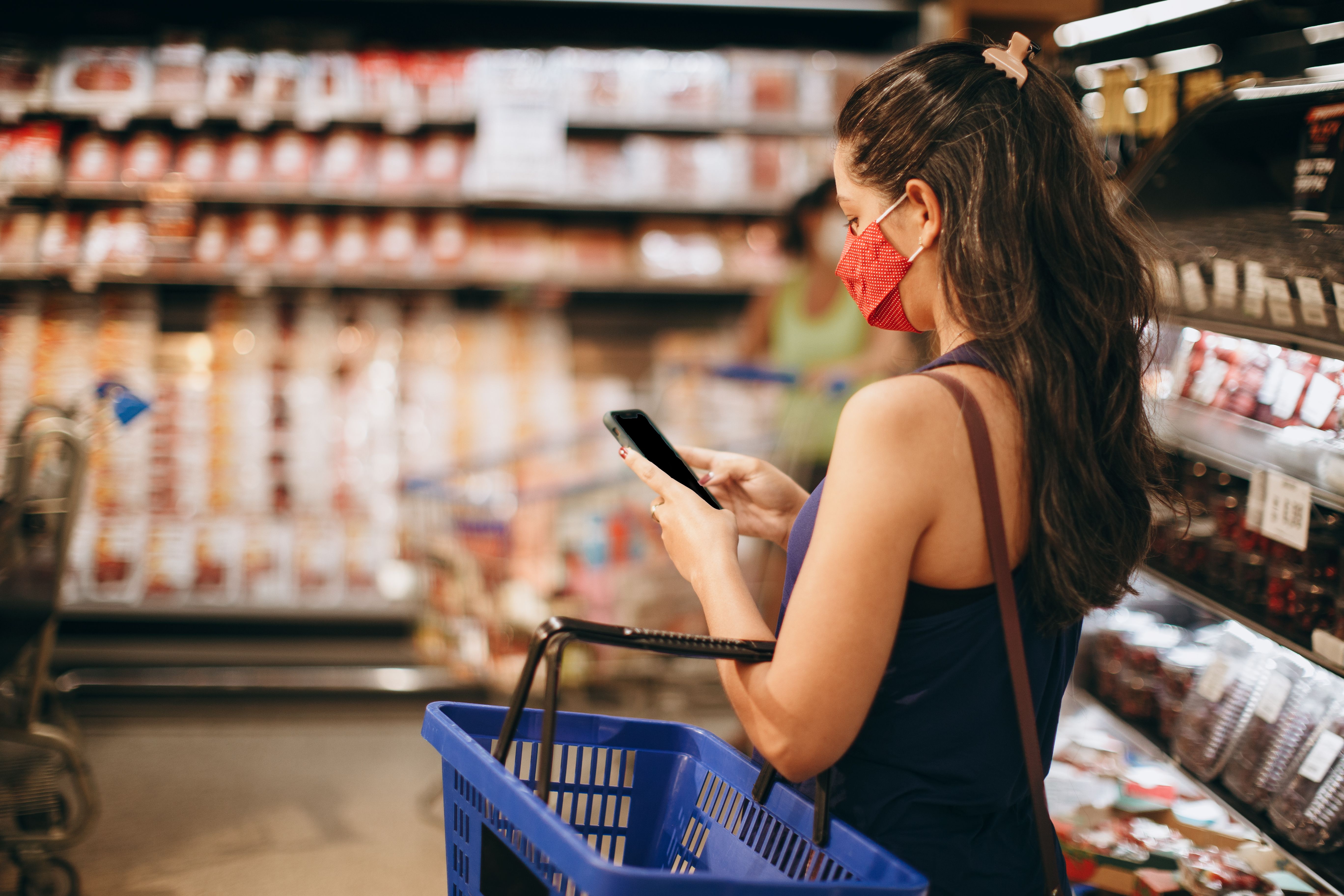 Side view of a woman wearing a red face mask using a smartphone while holding a blue shopping basket in a grocery aisle.