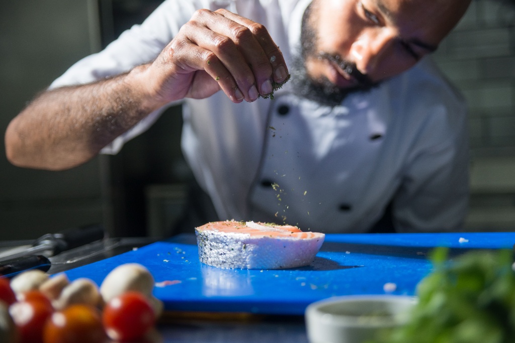 Close-up of a chef seasoning a fresh piece of salmon on a blue cutting board in a professional kitchen.