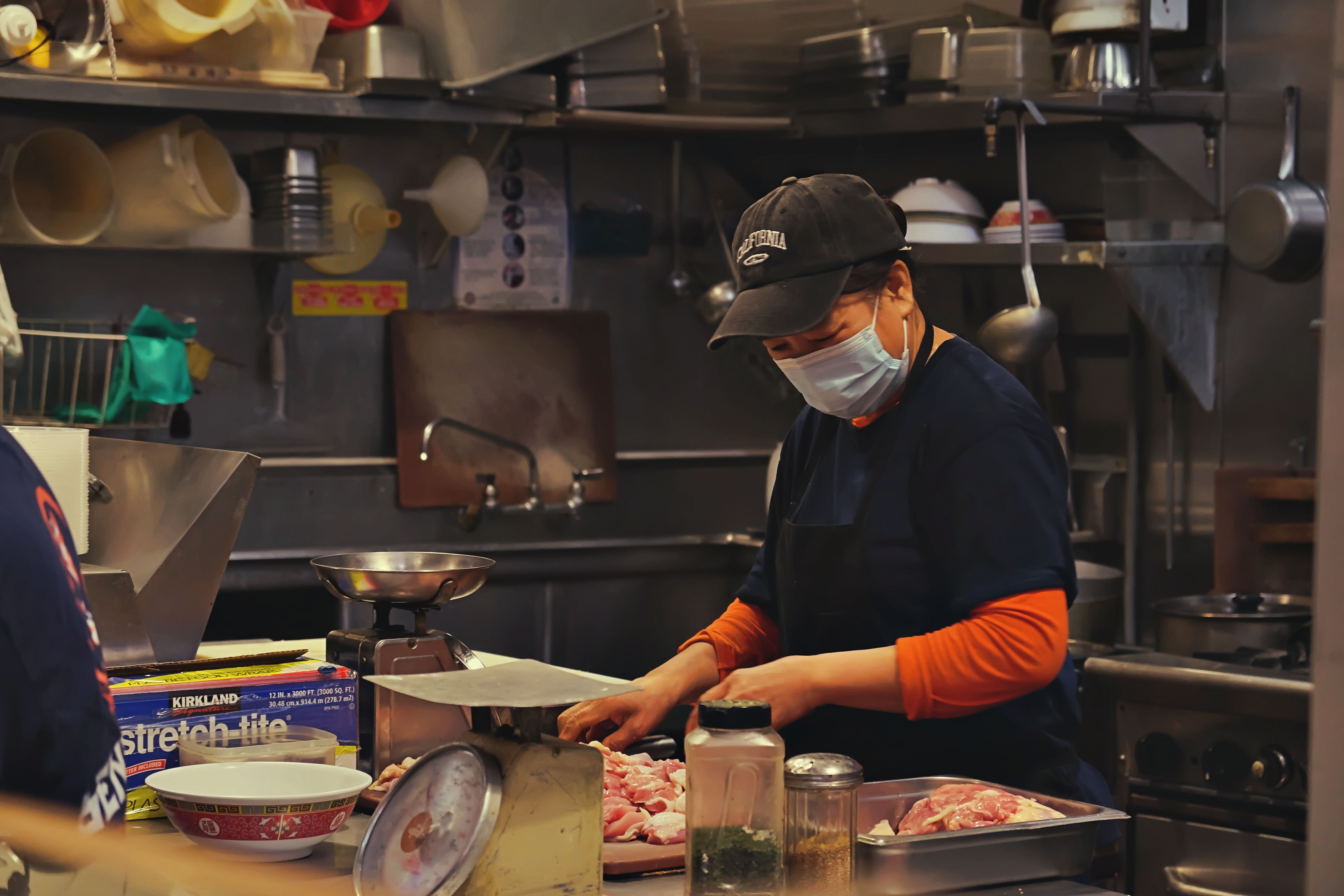 A kitchen worker wearing a black cap, face mask, and apron prepping raw meat on a cutting board near a weighing scale and food storage containers.