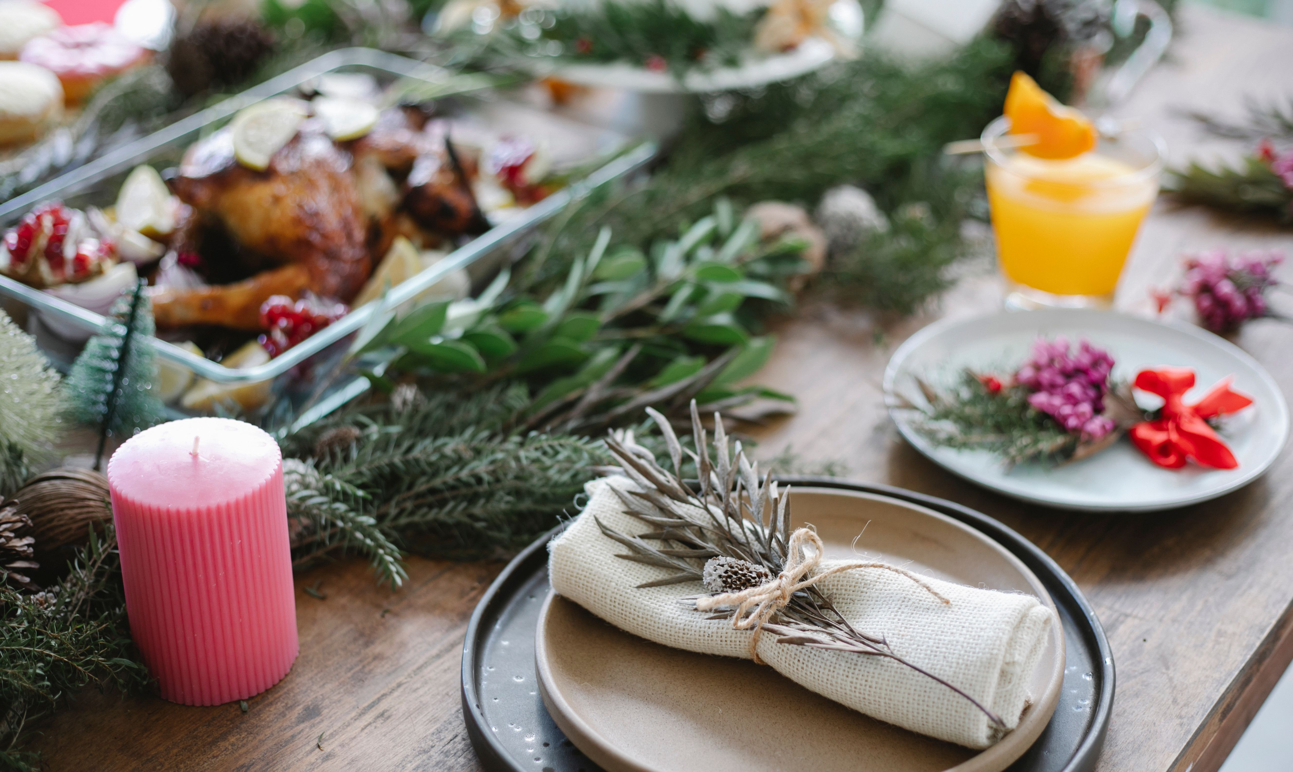 A festive Christmas dinner table setting featuring a roasted bird, evergreen garlands, a pink candle, and a wrapped napkin with a dried branch.