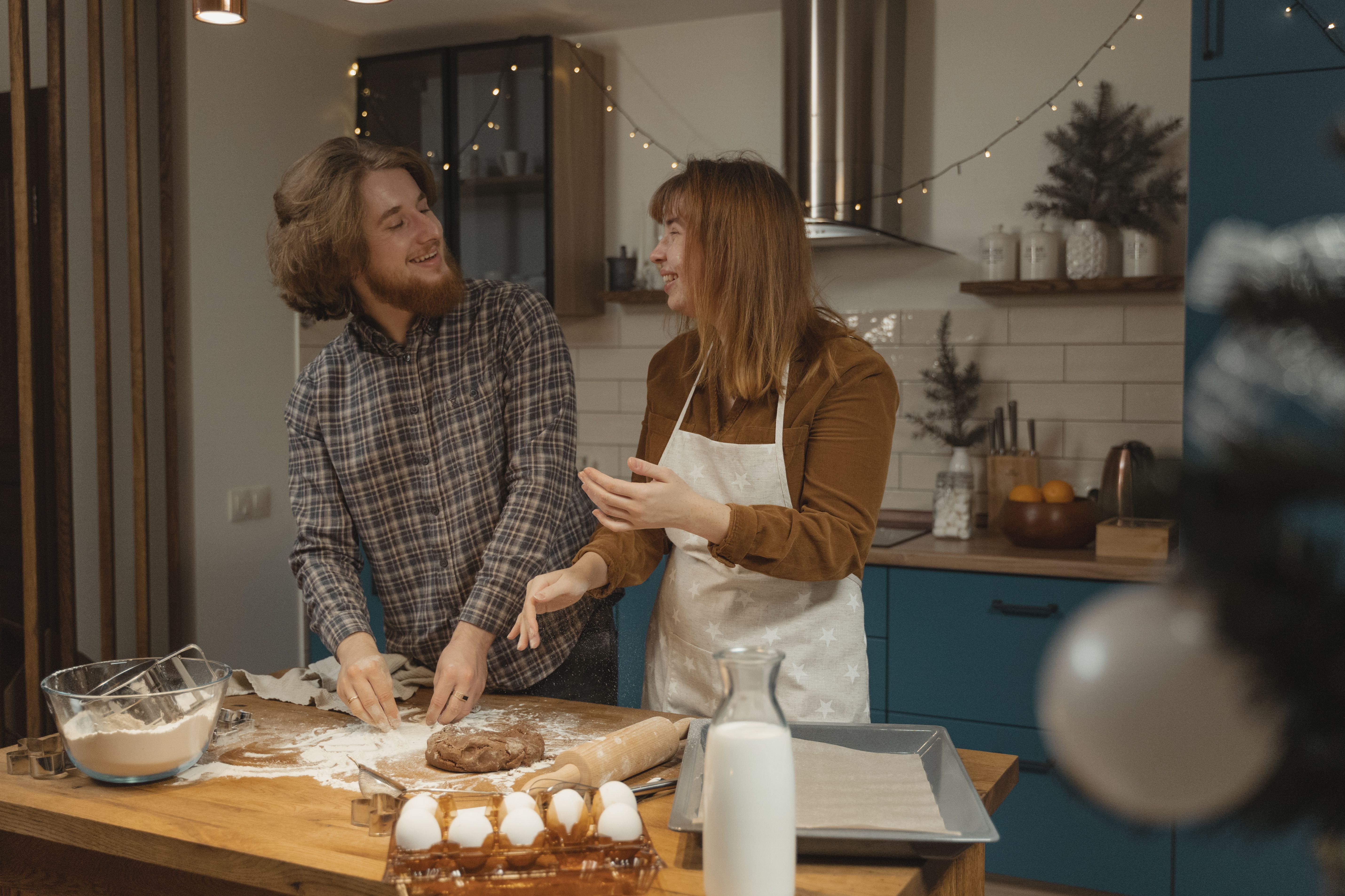 A smiling man and woman wearing an apron baking together at a wooden kitchen counter covered in flour, gingerbread dough, and eggs.
