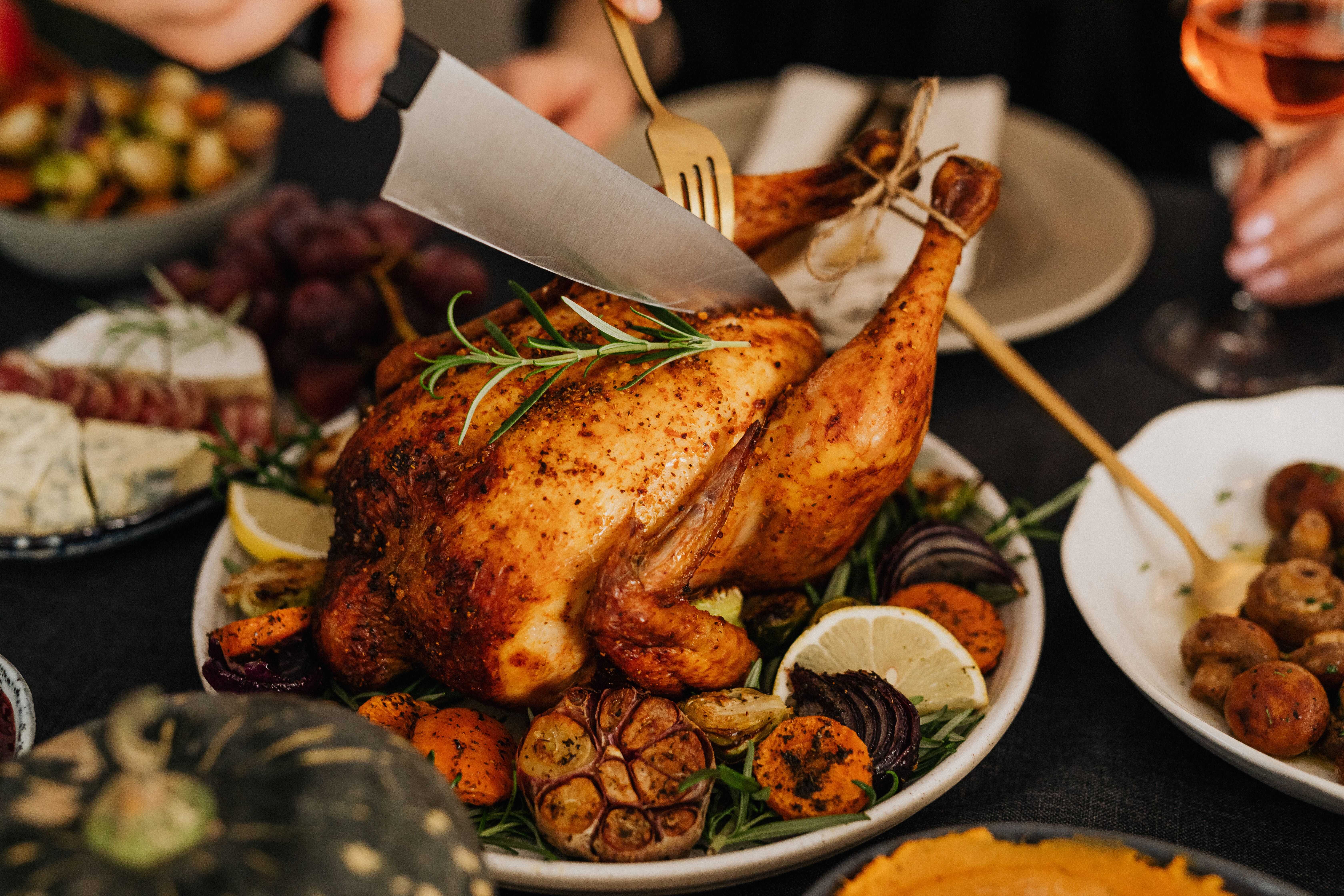 lose-up of hands carving a whole roasted chicken garnished with fresh rosemary, lemons, and roasted vegetables.