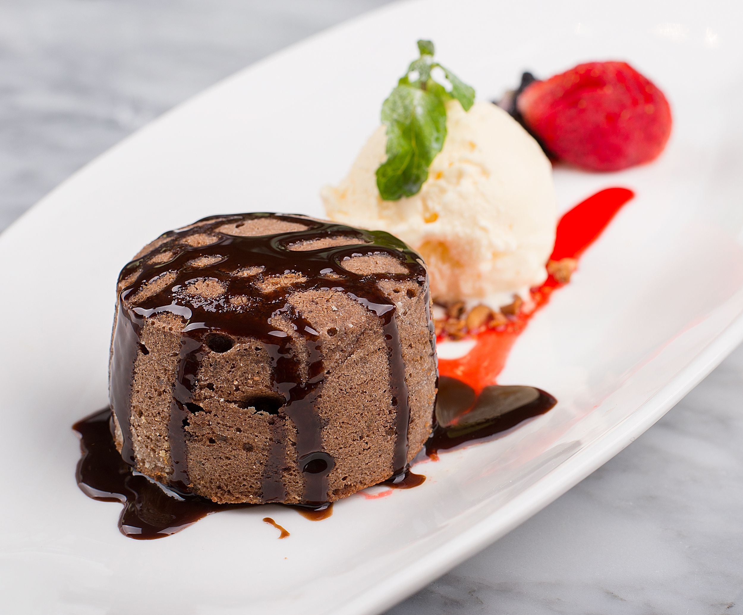 A chocolate lava cake drizzled with chocolate syrup served alongside a scoop of vanilla ice cream and a fresh strawberry on a white plate.