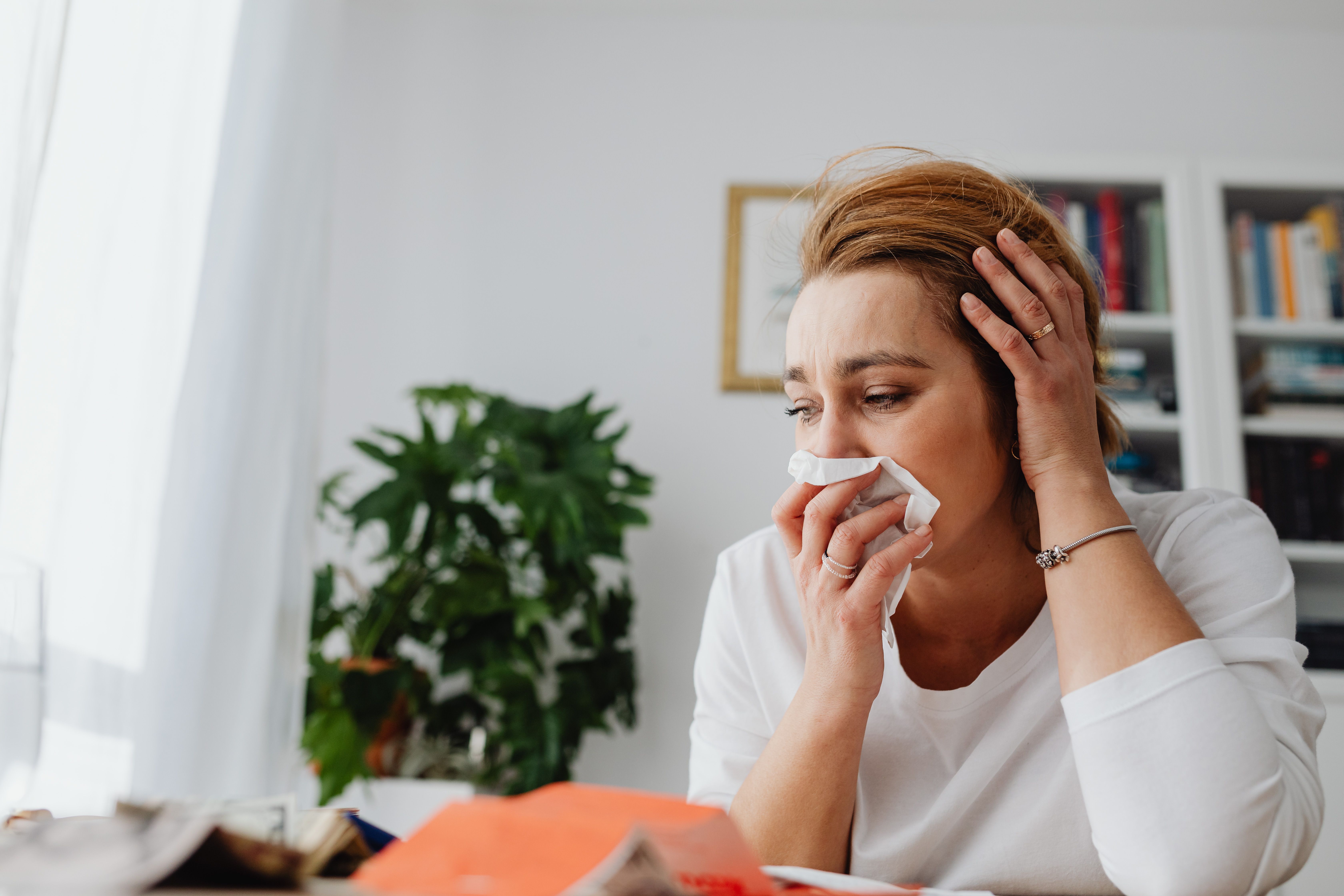 A distressed woman sitting at a desk holding a tissue to her face with one hand on her head.