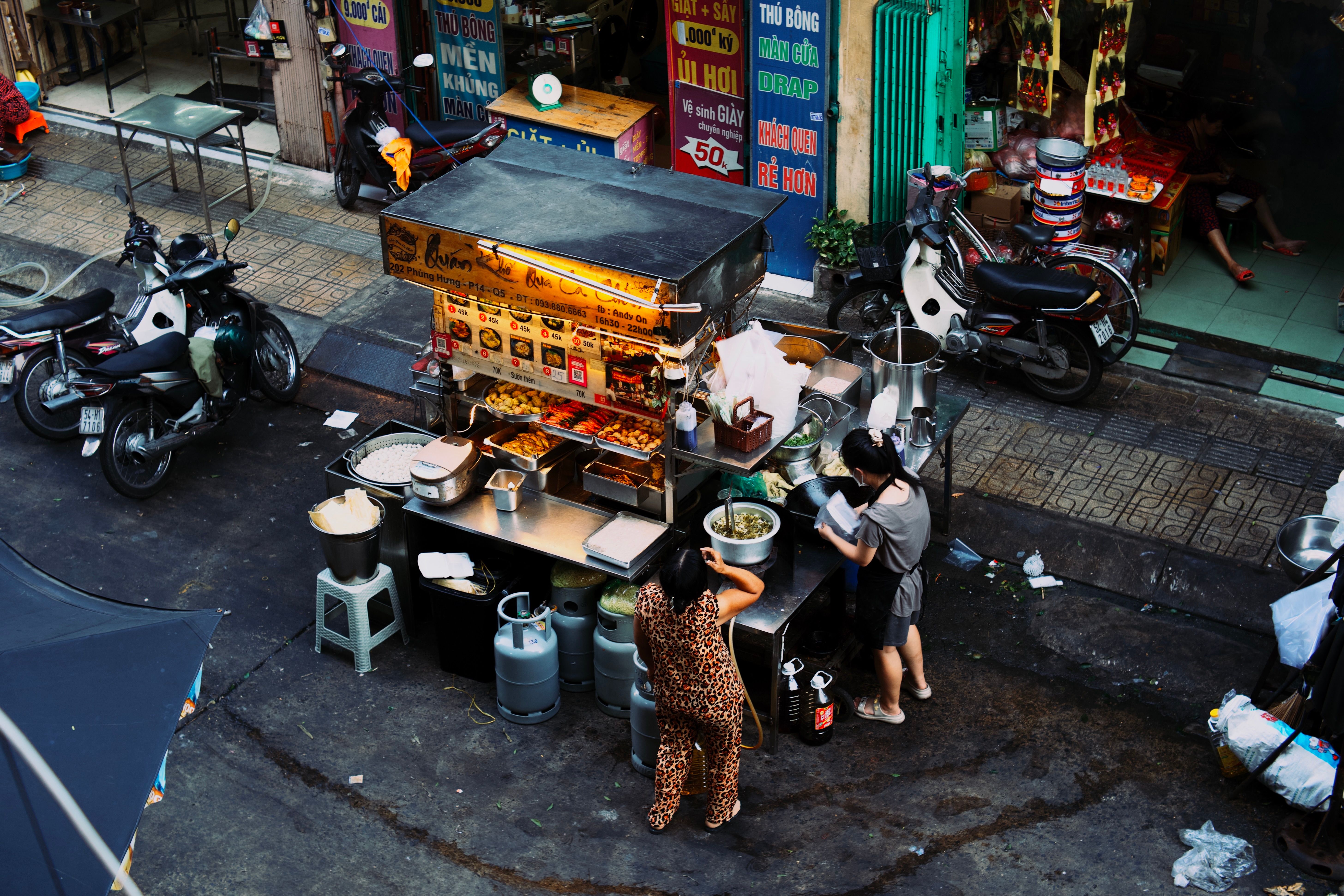 High-angle view of a street food stall in an urban alleyway surrounded by parked motorbikes and workers preparing food.