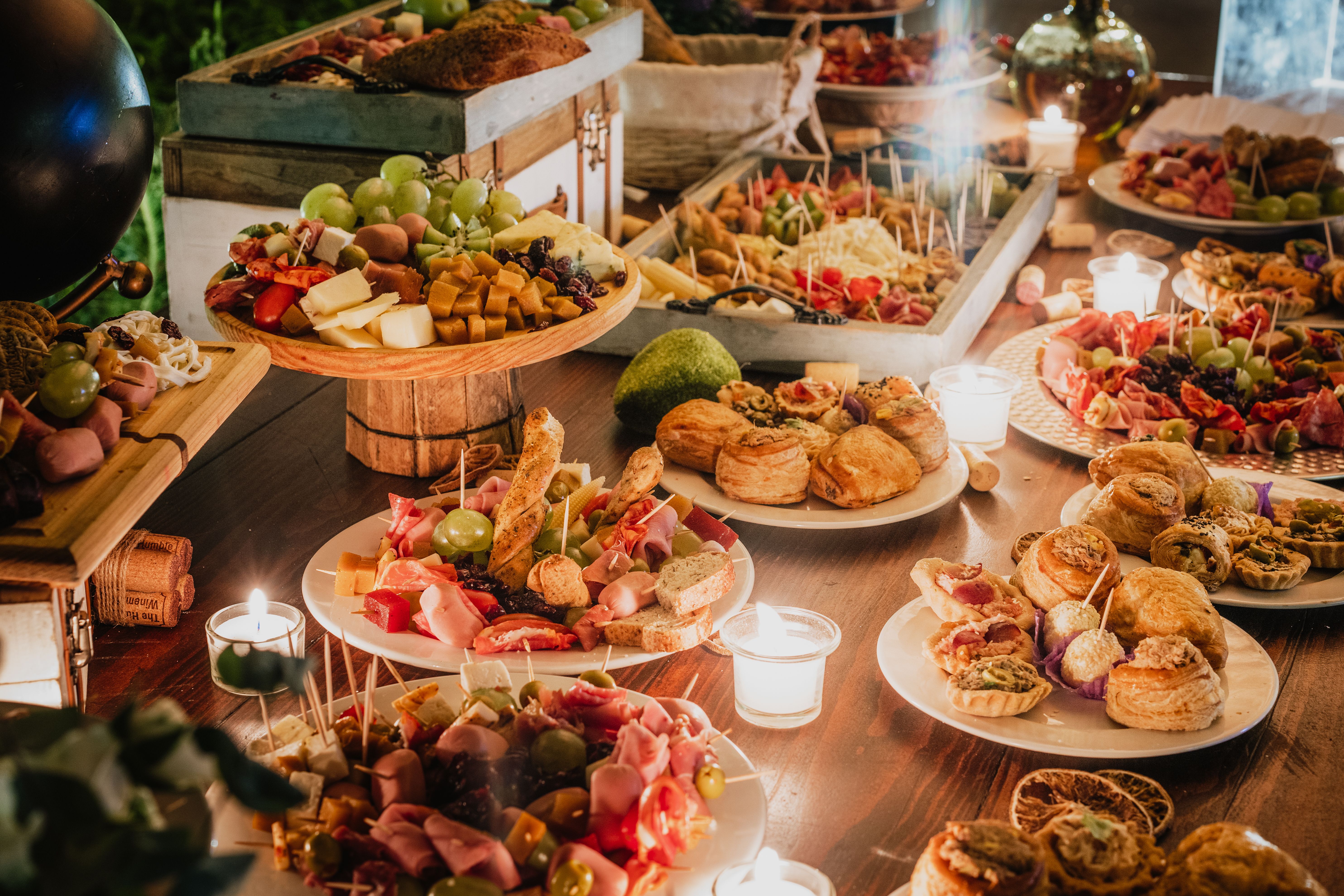 An extensive buffet table spread featuring charcuterie boards, cheeses, fruits, and small baked pastries lit by candles.
