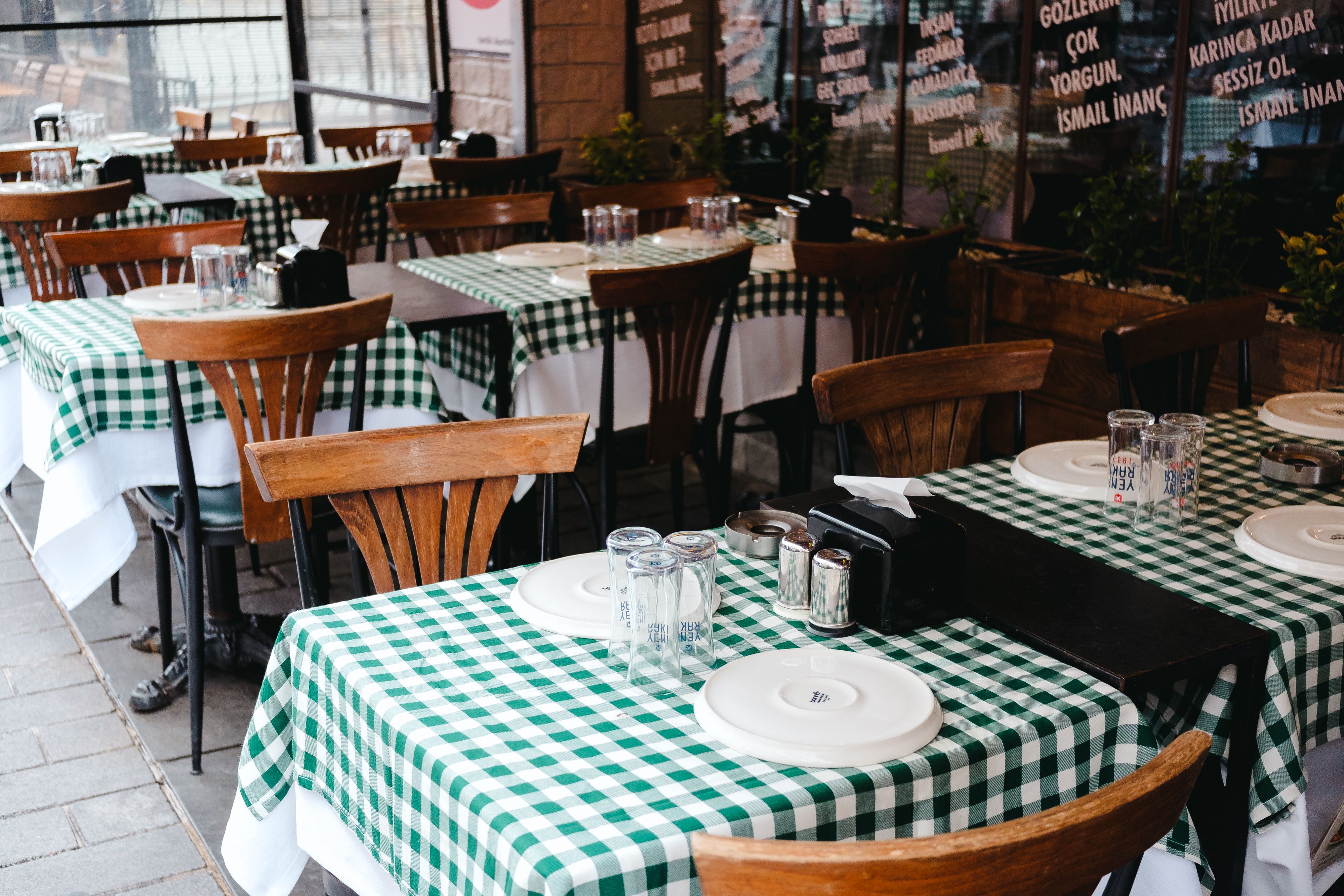 An empty outdoor restaurant seating area with green and white checkered tablecloths and stacked glasses on the tables.