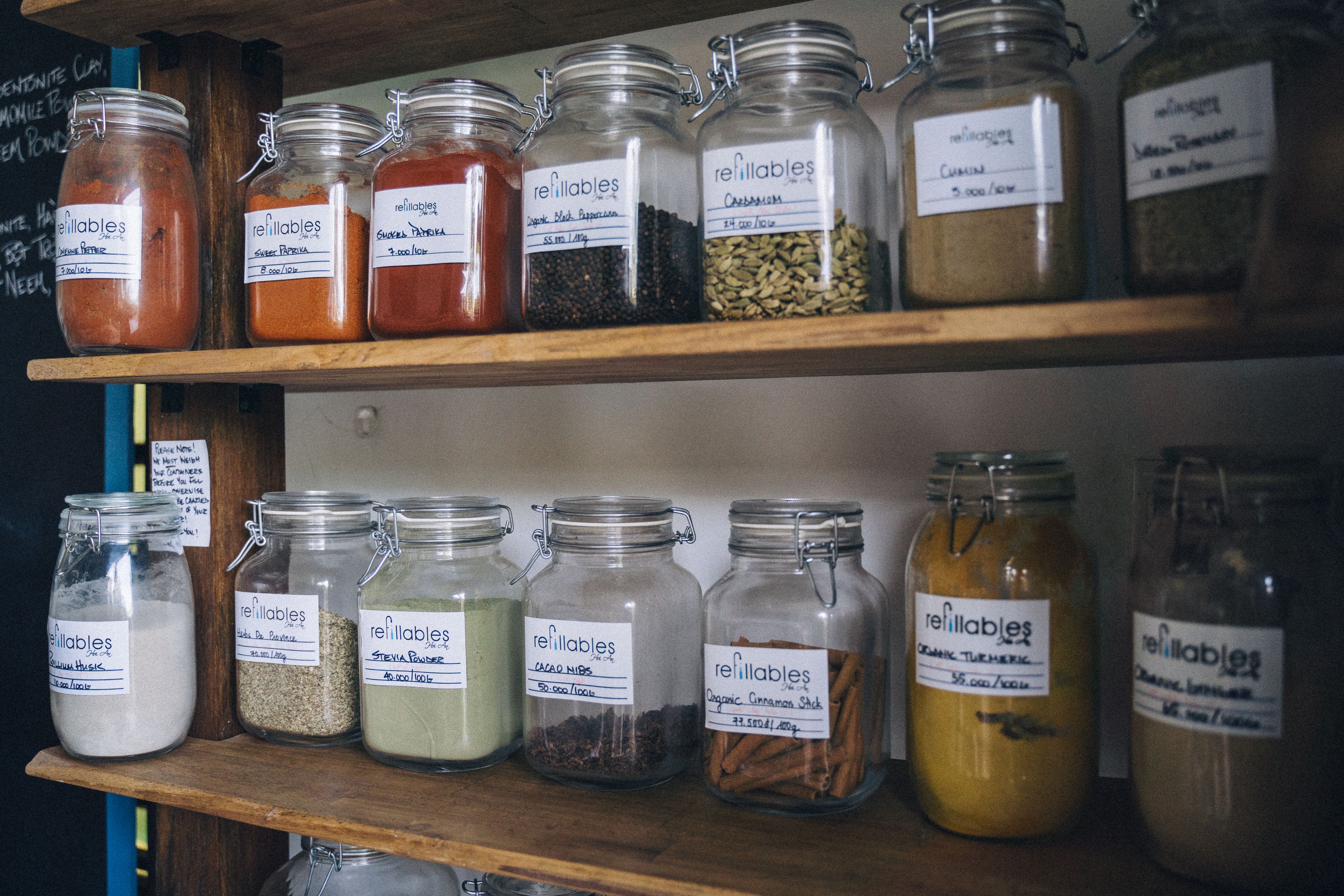 Wooden shelves stocked with labeled glass jars containing various spices, powders, and dried herbs in a zero-waste refillery store.