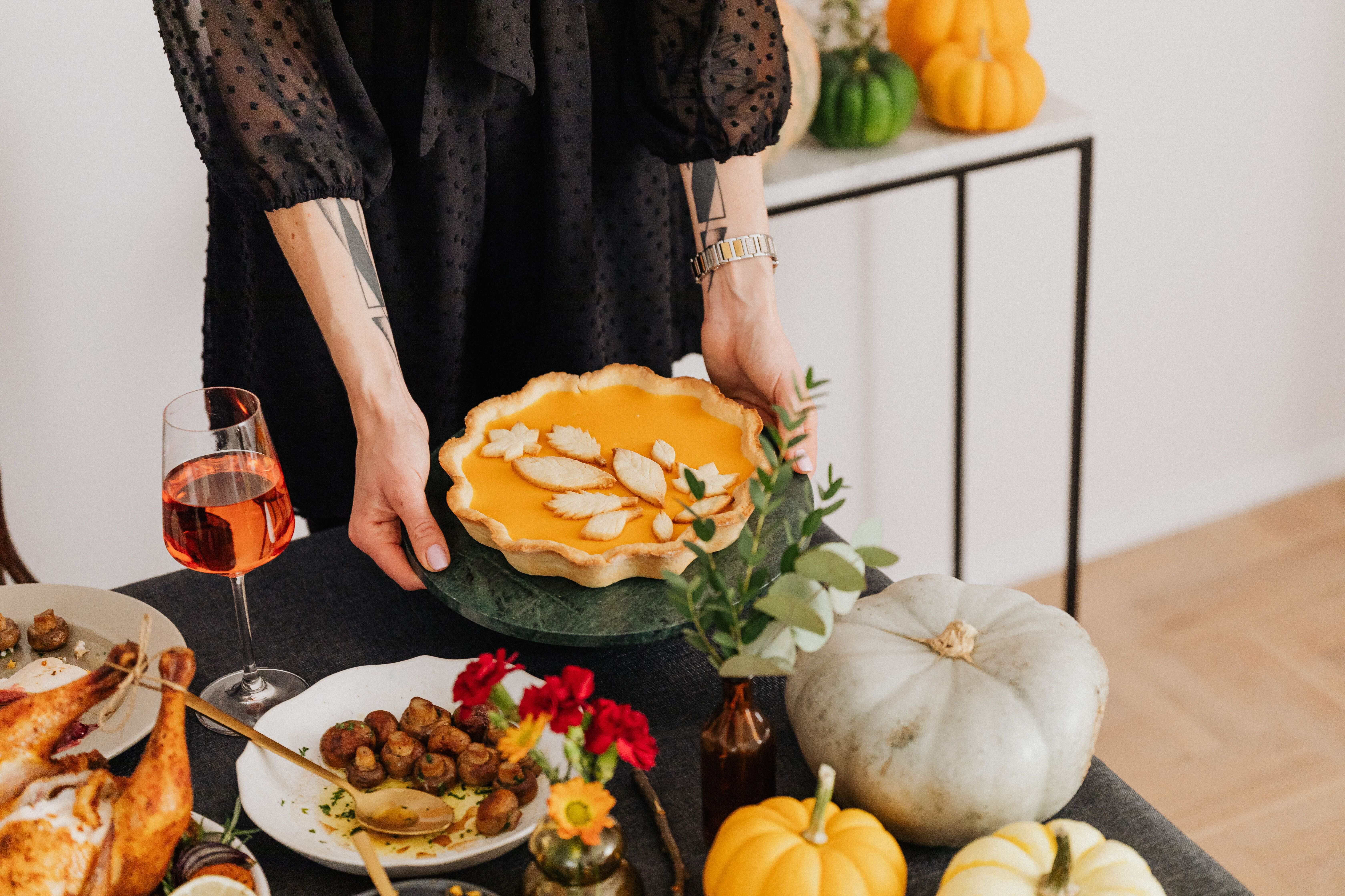 A person holding a baked pumpkin pie decorated with pastry leaves over a autumn dinner table setup with gourds and wine.