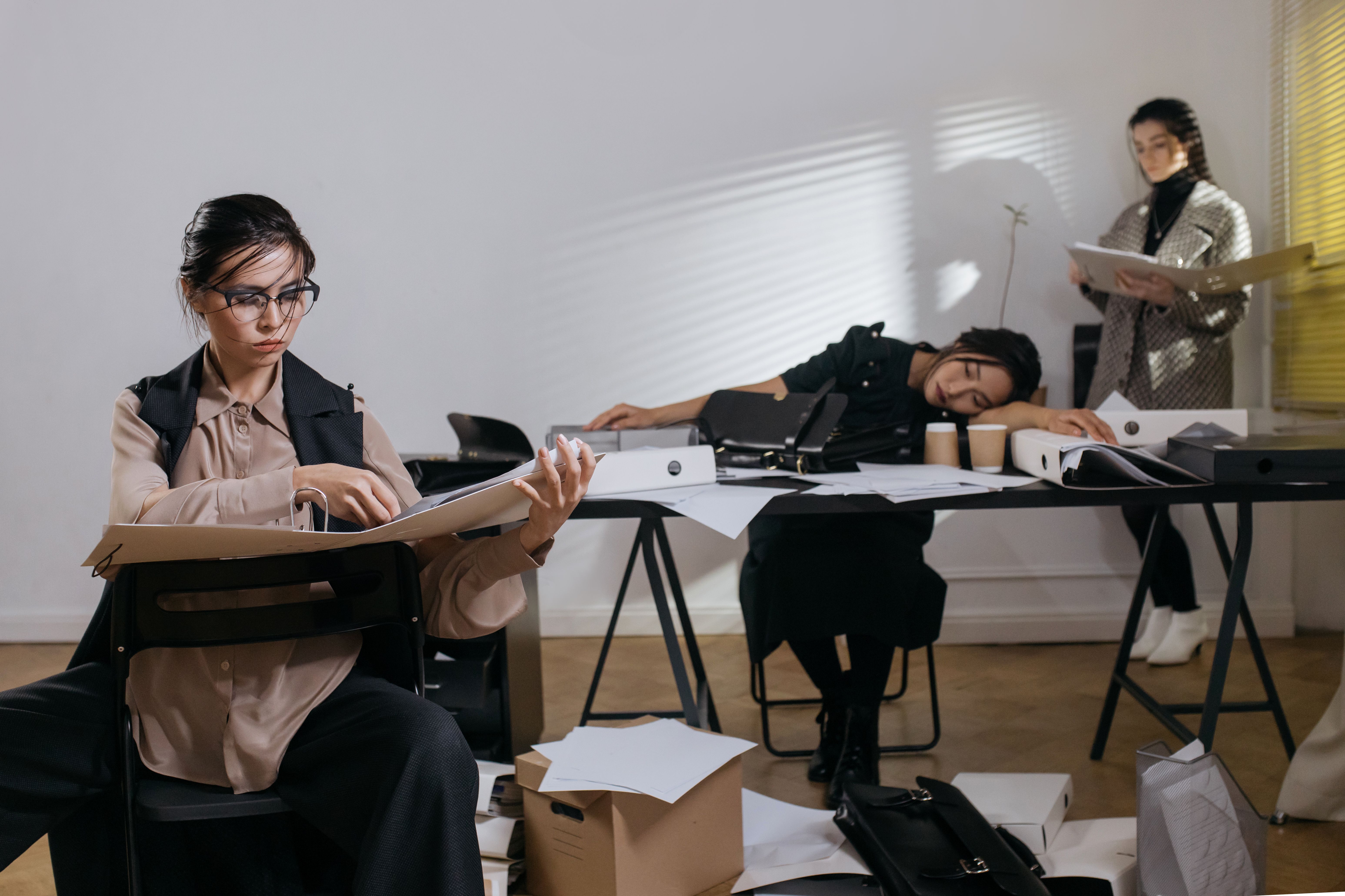 An exhausted office team overwhelmed with paperwork, including a woman sitting with a binder surrounded by cluttered documents and another sleeping at her desk.