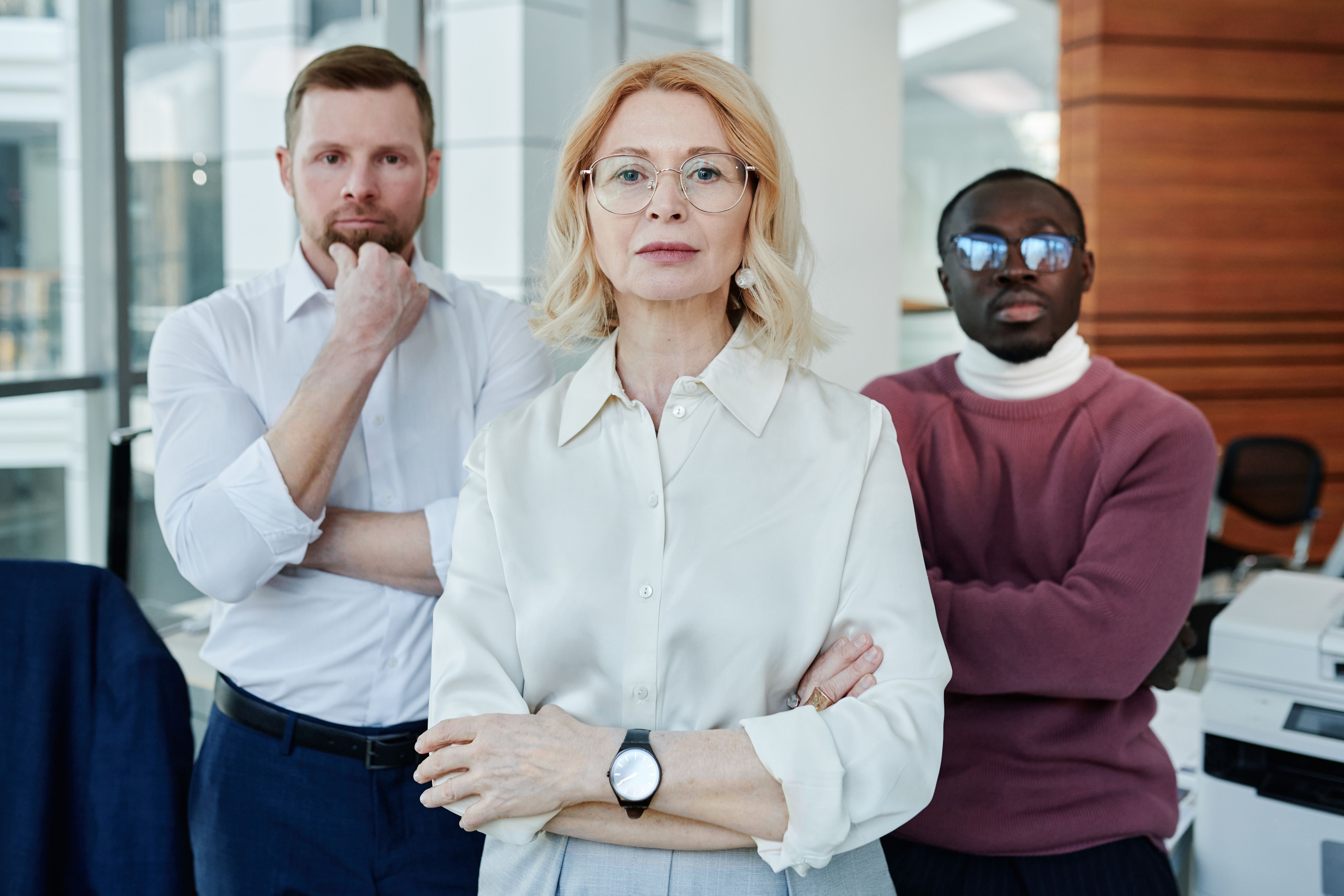 Three corporate professionals standing side by side with confident expressions in a modern office setting.