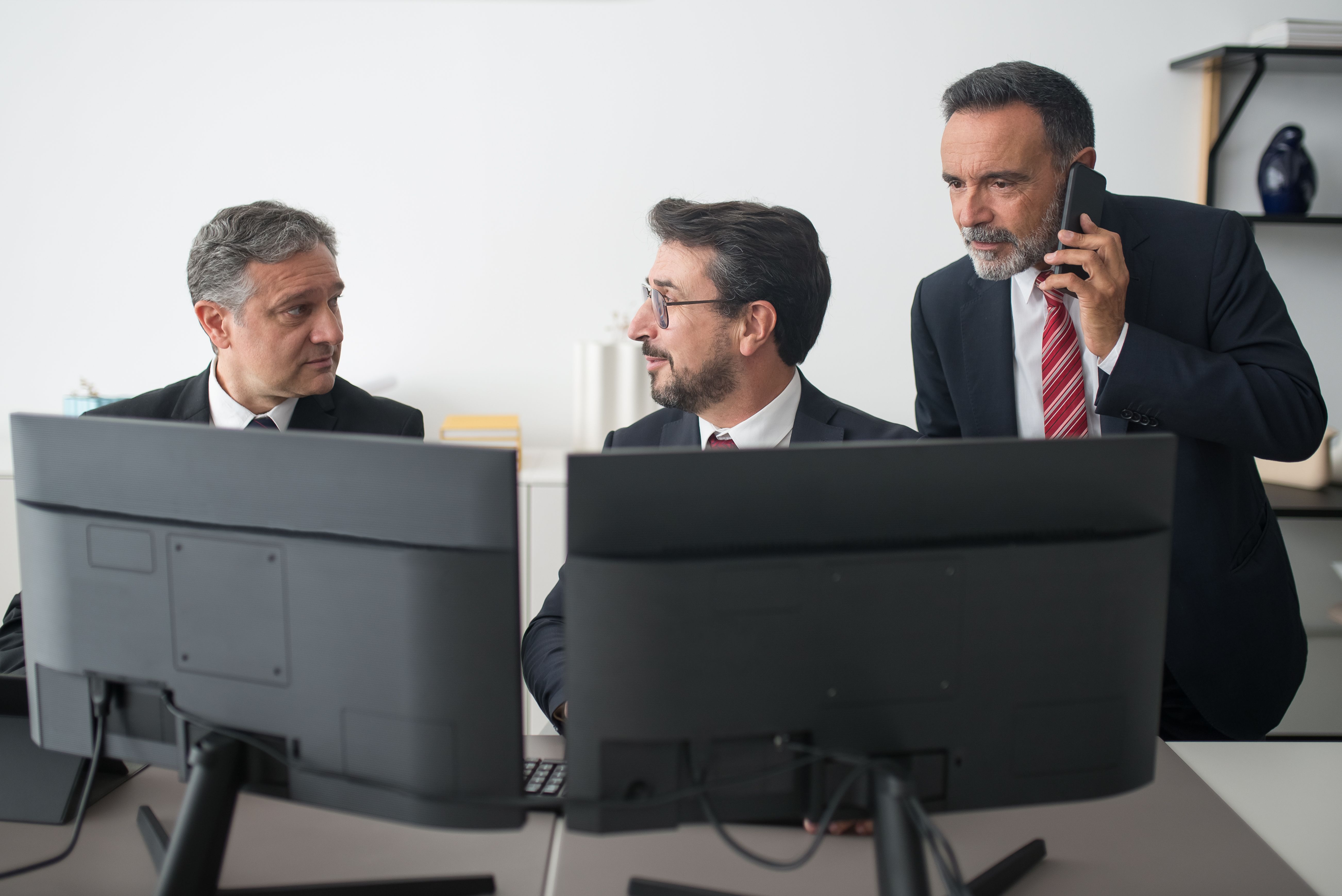 Three businessmen in suits discussing work behind dual computer monitors while one makes a phone call.