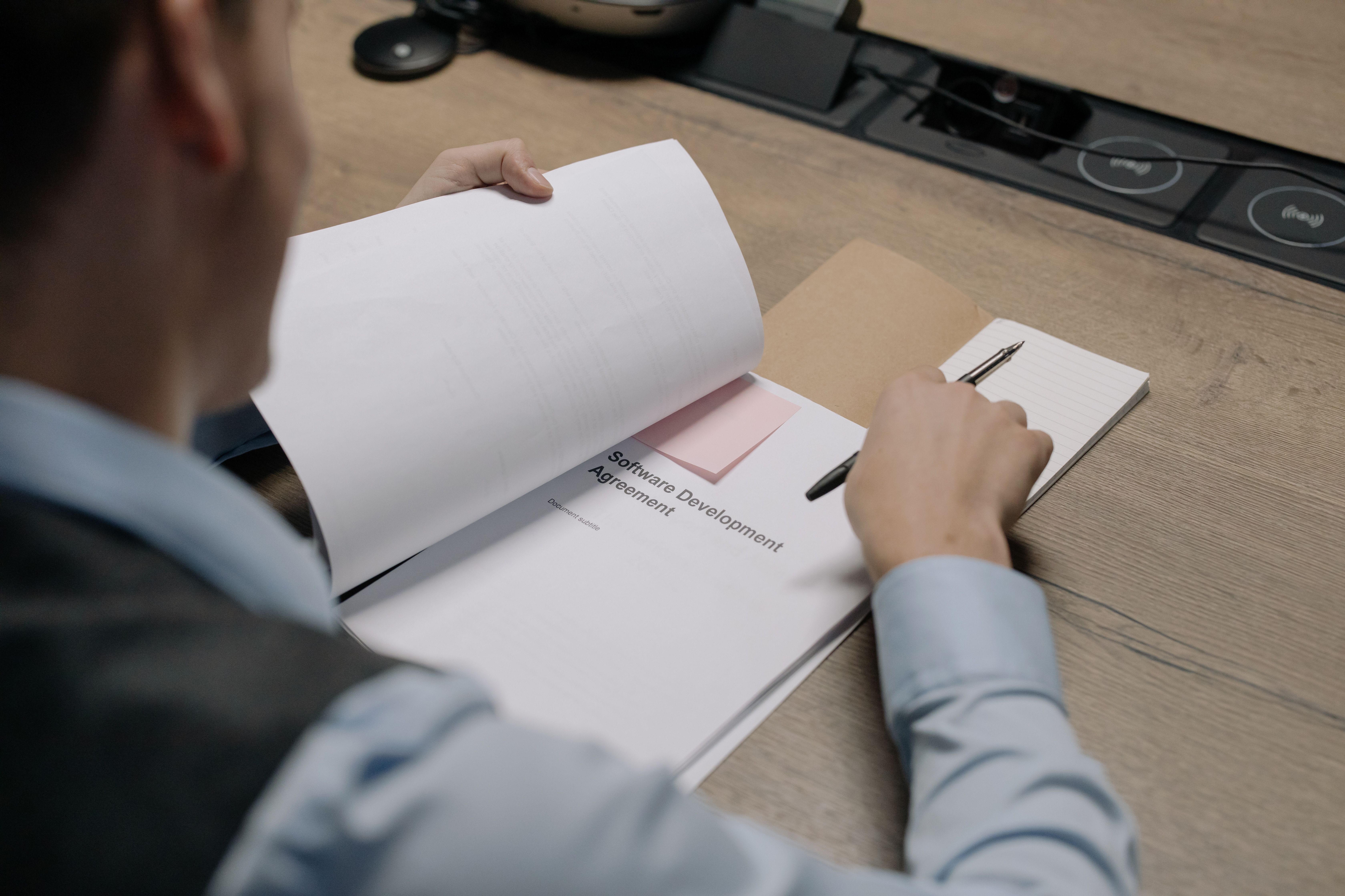 A person at a wooden desk reviewing a printed document titled Software Development Agreement and writing in a notepad with a pen.