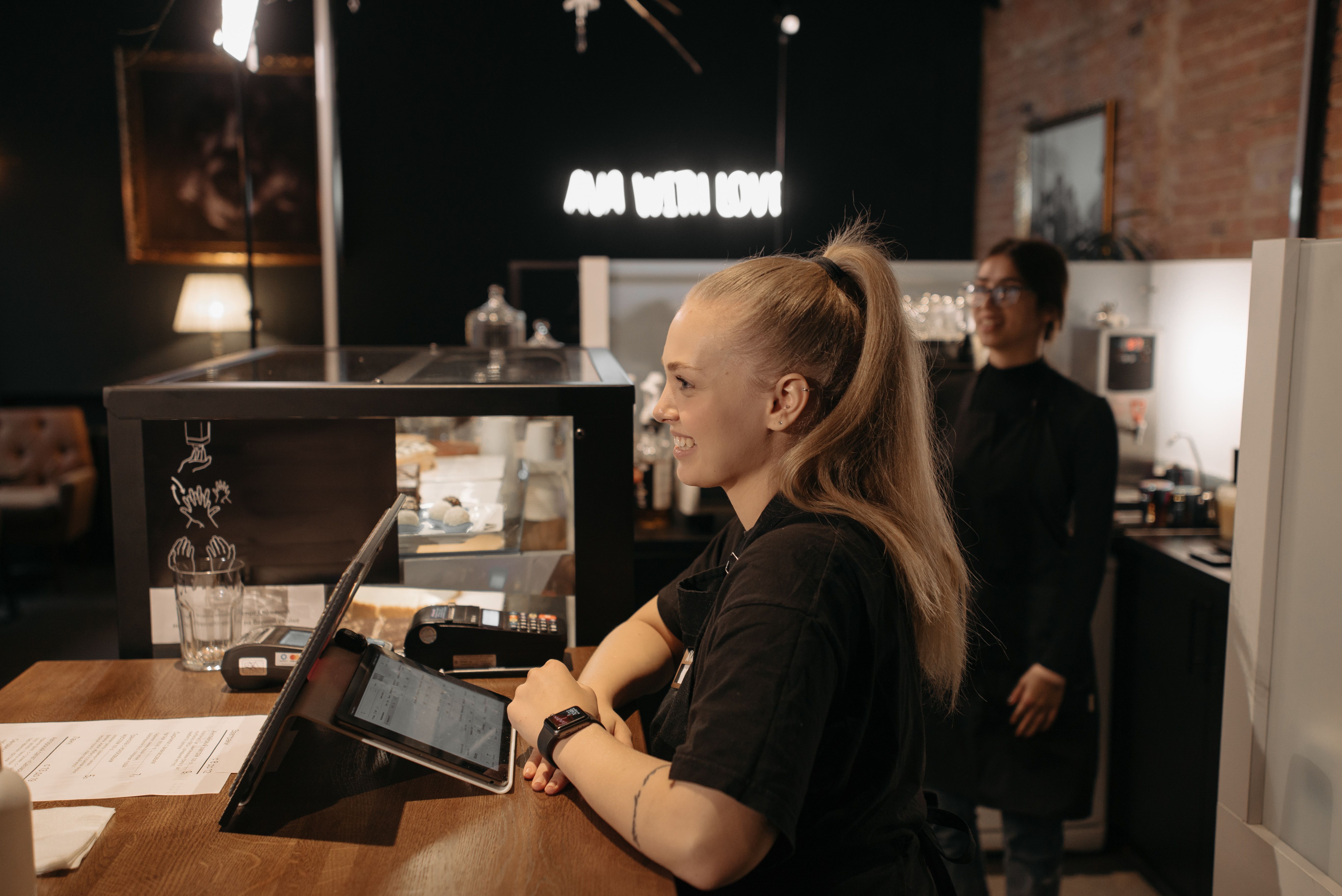 A blonde cafe employee smiling while using a point-of-sale tablet system on a wooden counter.
