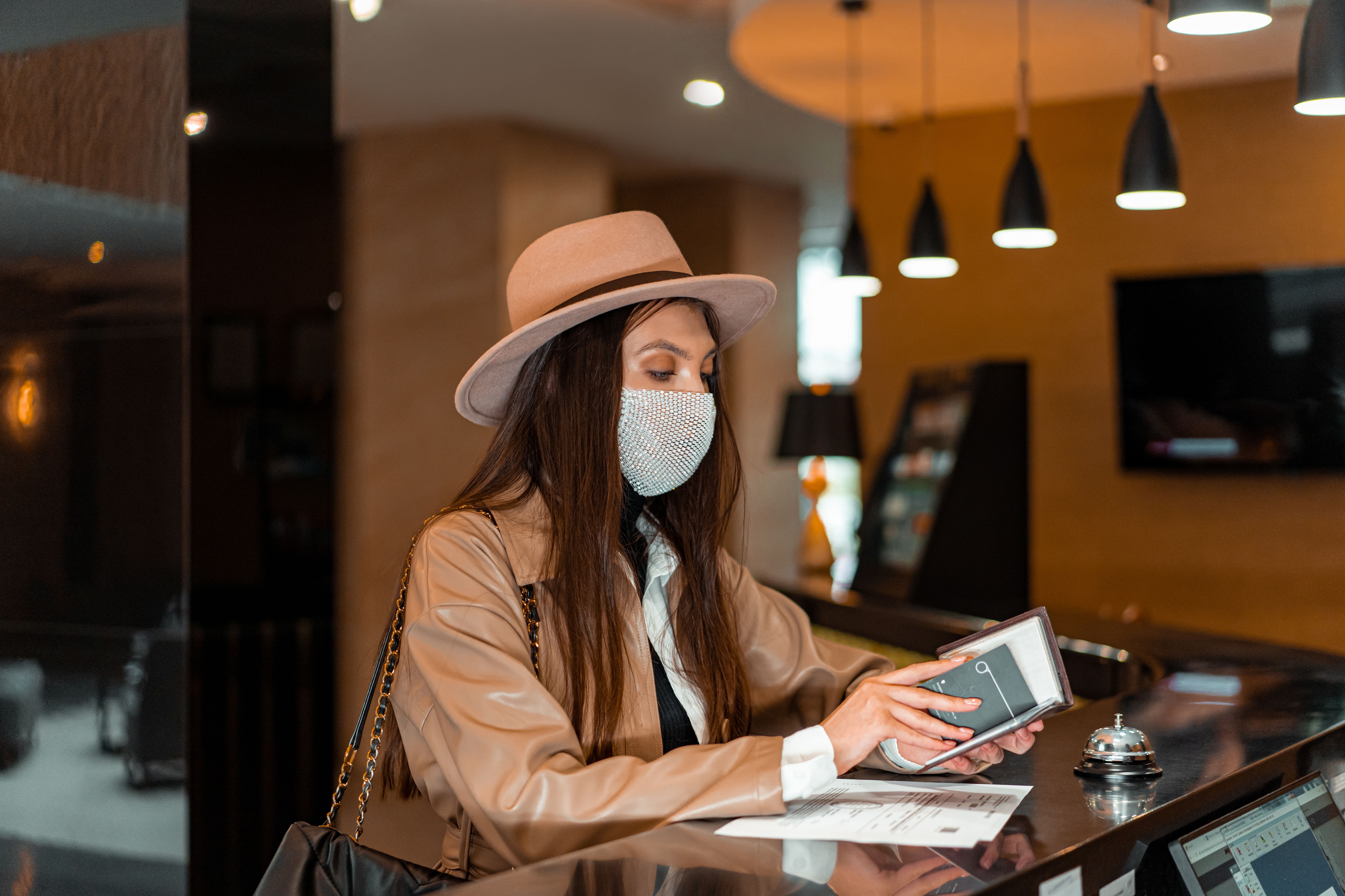 A woman wearing a brown hat and a sparkling face mask checking in at a hotel reception desk holding a wallet or document folder.