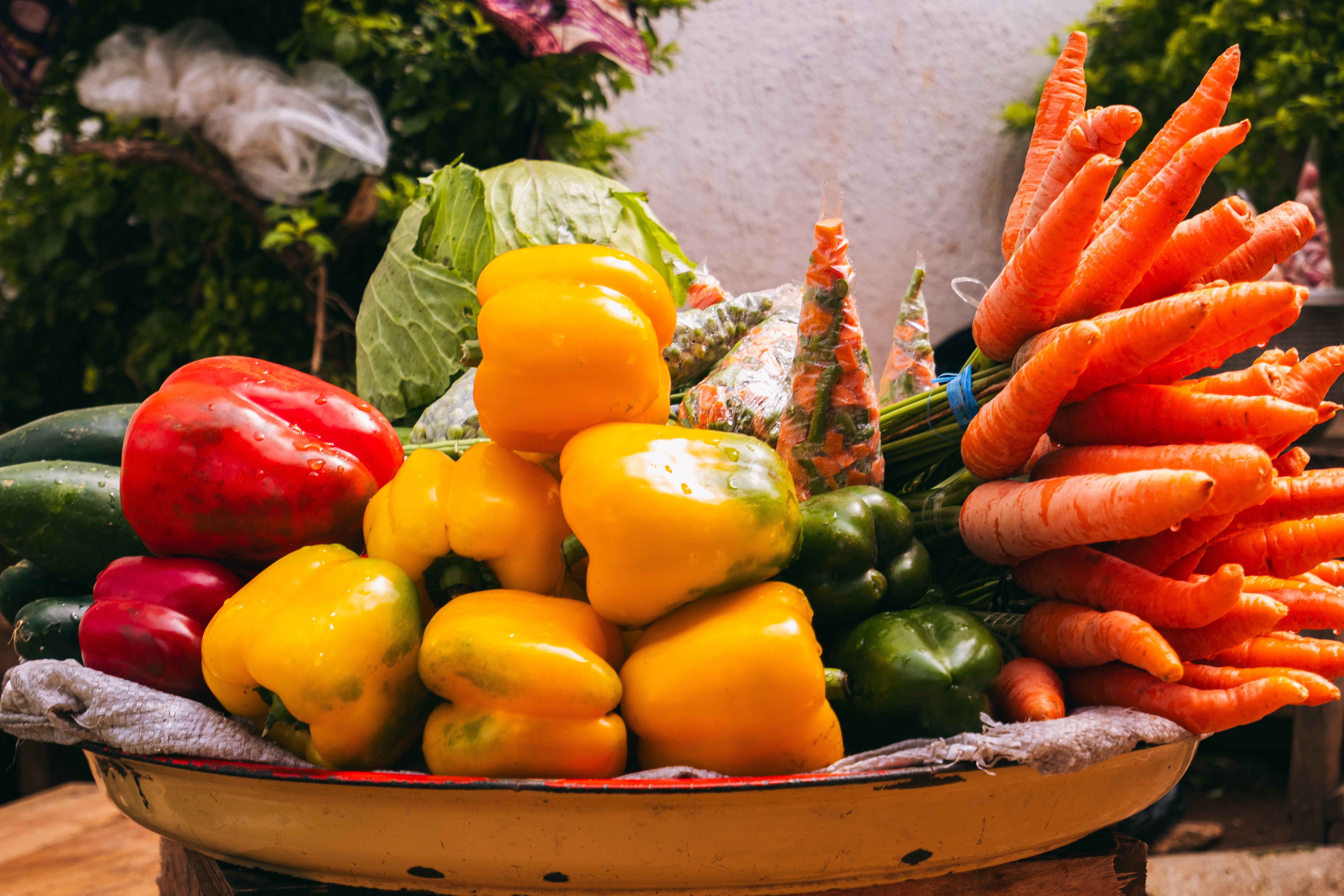 A large platter filled with colorful fresh vegetables, including bright yellow and red bell peppers, green cucumbers, and a bundle of fresh carrots.
