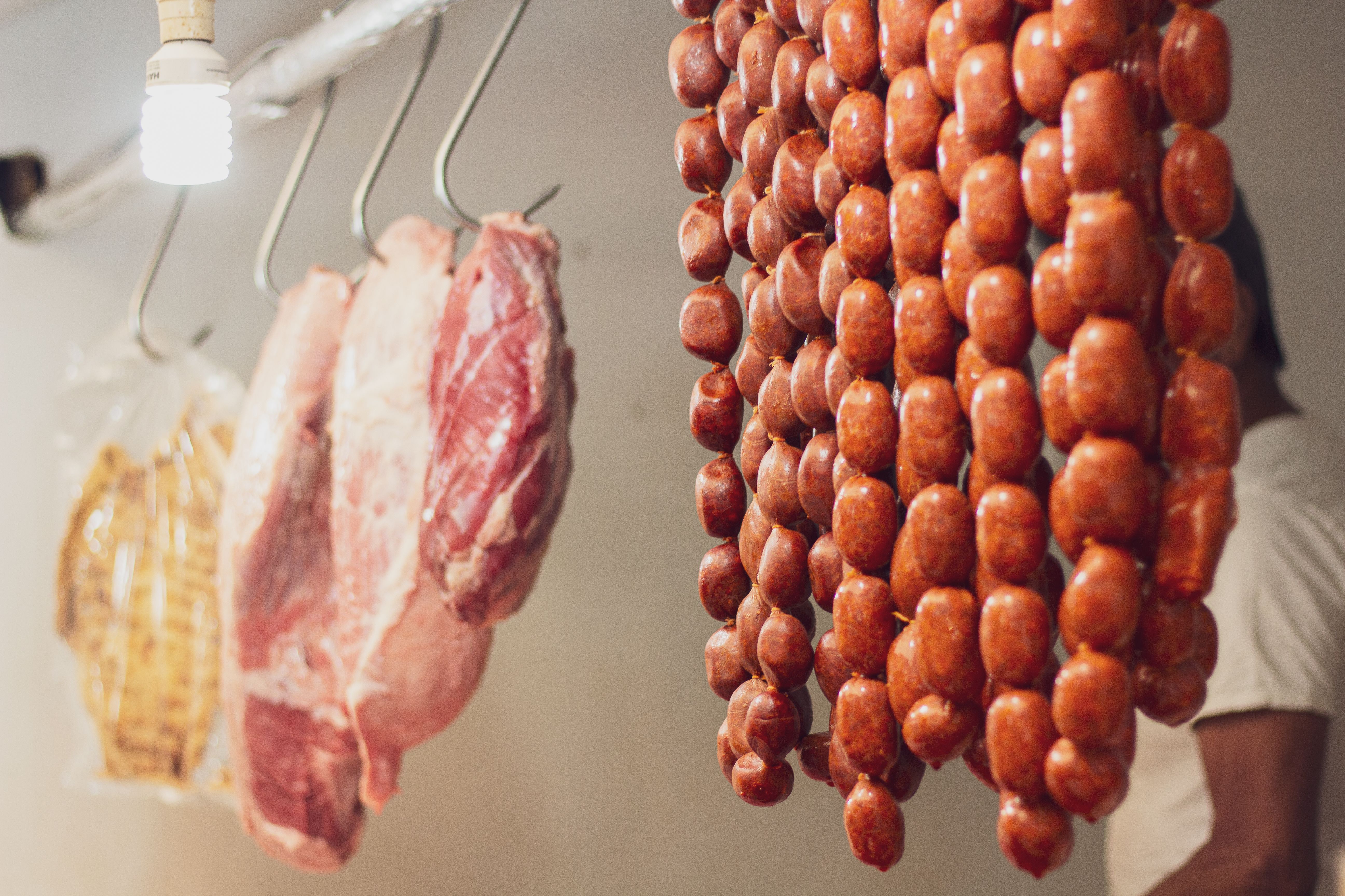 Strings of cured sausages hanging vertically alongside raw cuts of meat suspended from metal hooks under a bare light bulb.