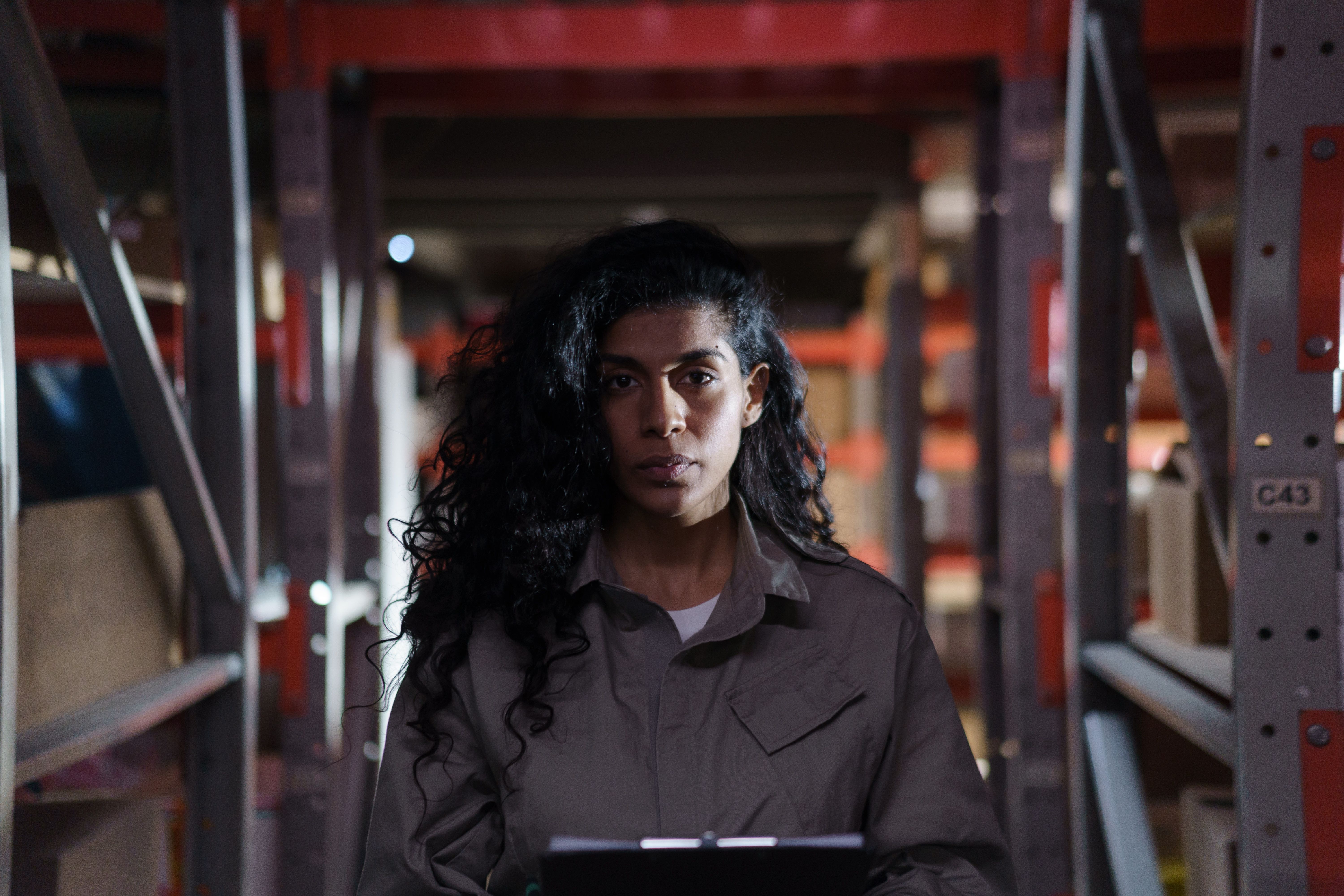 A woman with long curly hair in work coveralls holding a clipboard standing inside a distribution warehouse with storage racks.