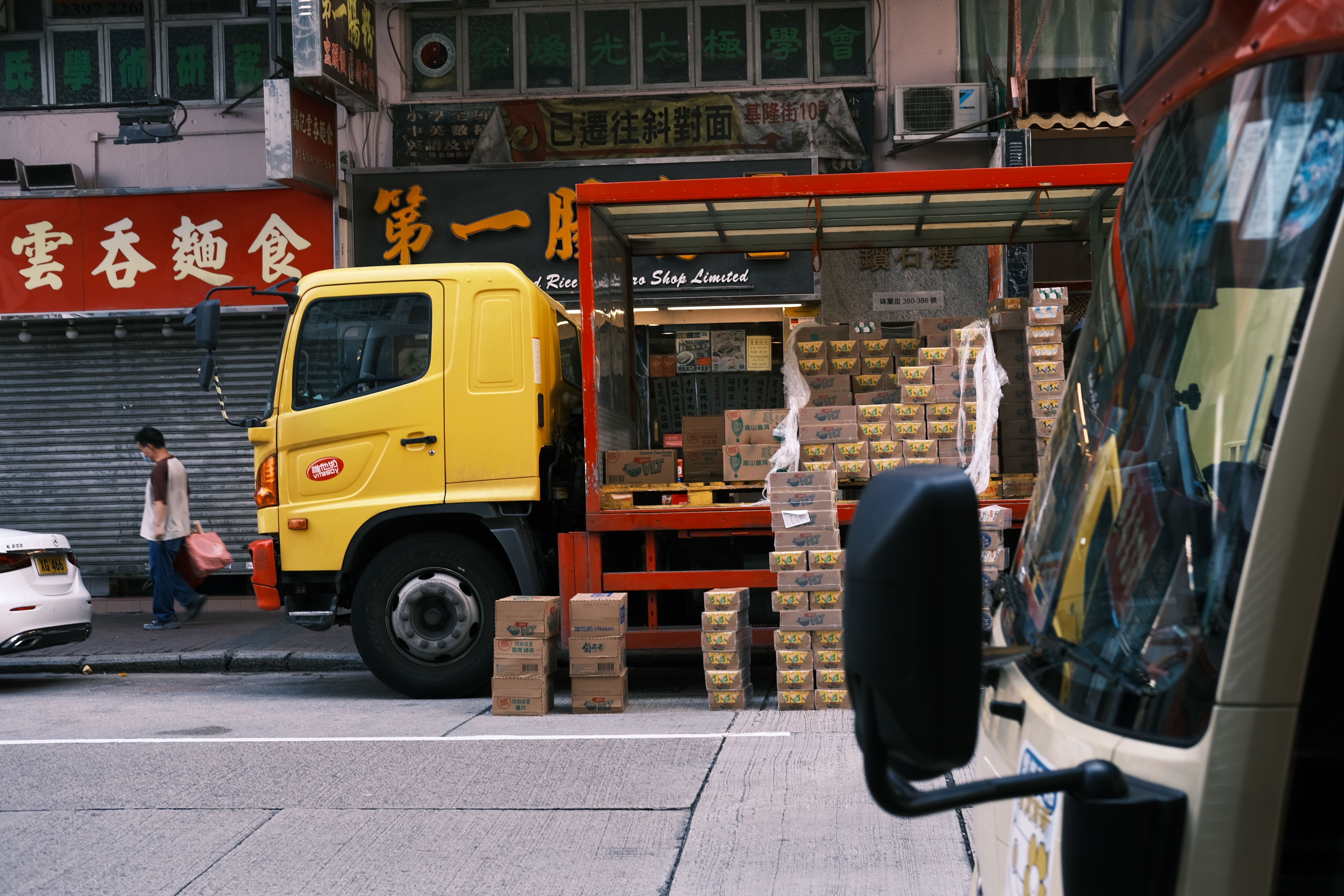 A yellow delivery truck parked on an urban street with its back open, filled with stacked cardboard boxes near storefronts with Asian signage.