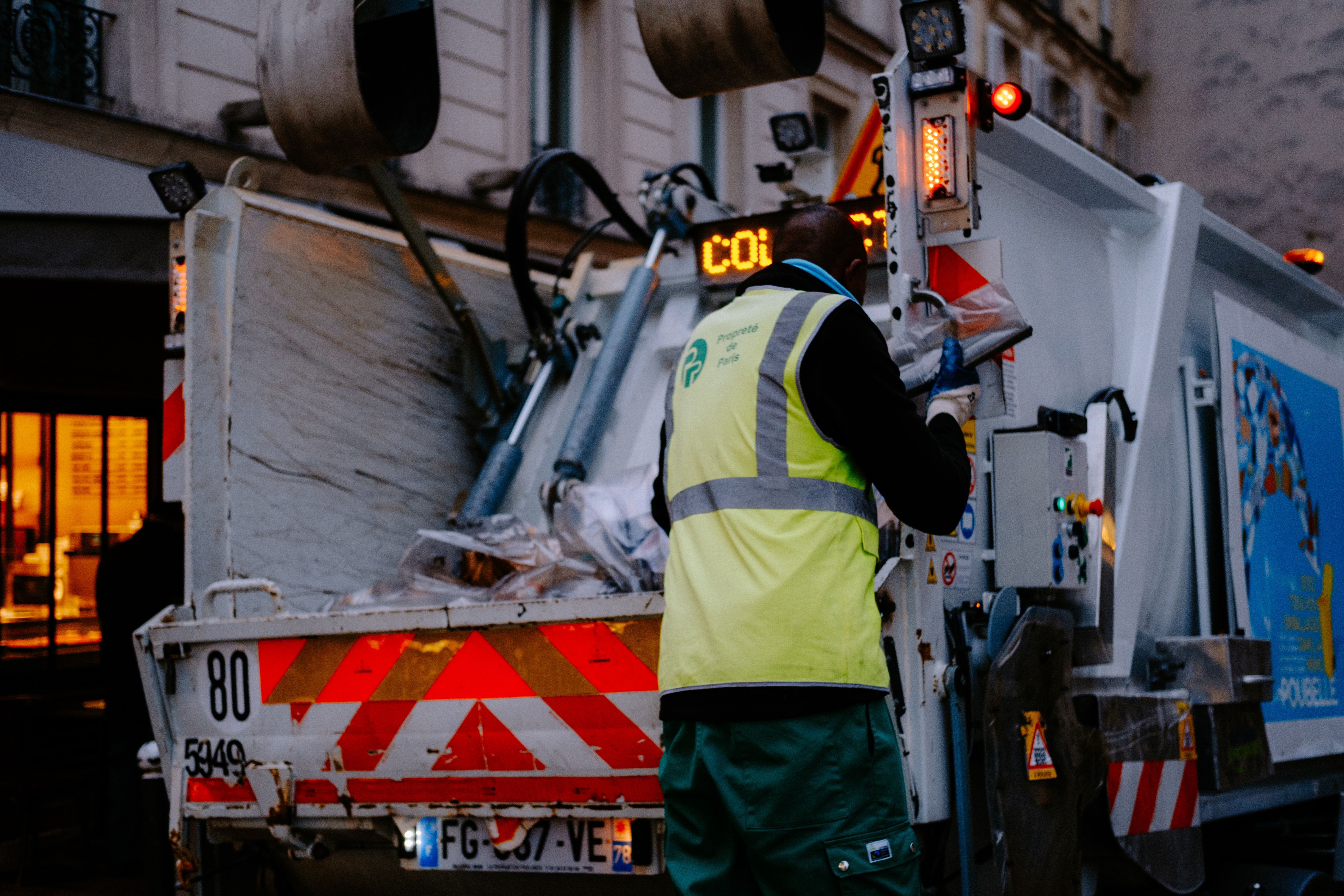 A sanitation worker in a high-visibility vest standing at the rear of a municipal garbage truck during evening waste collection.