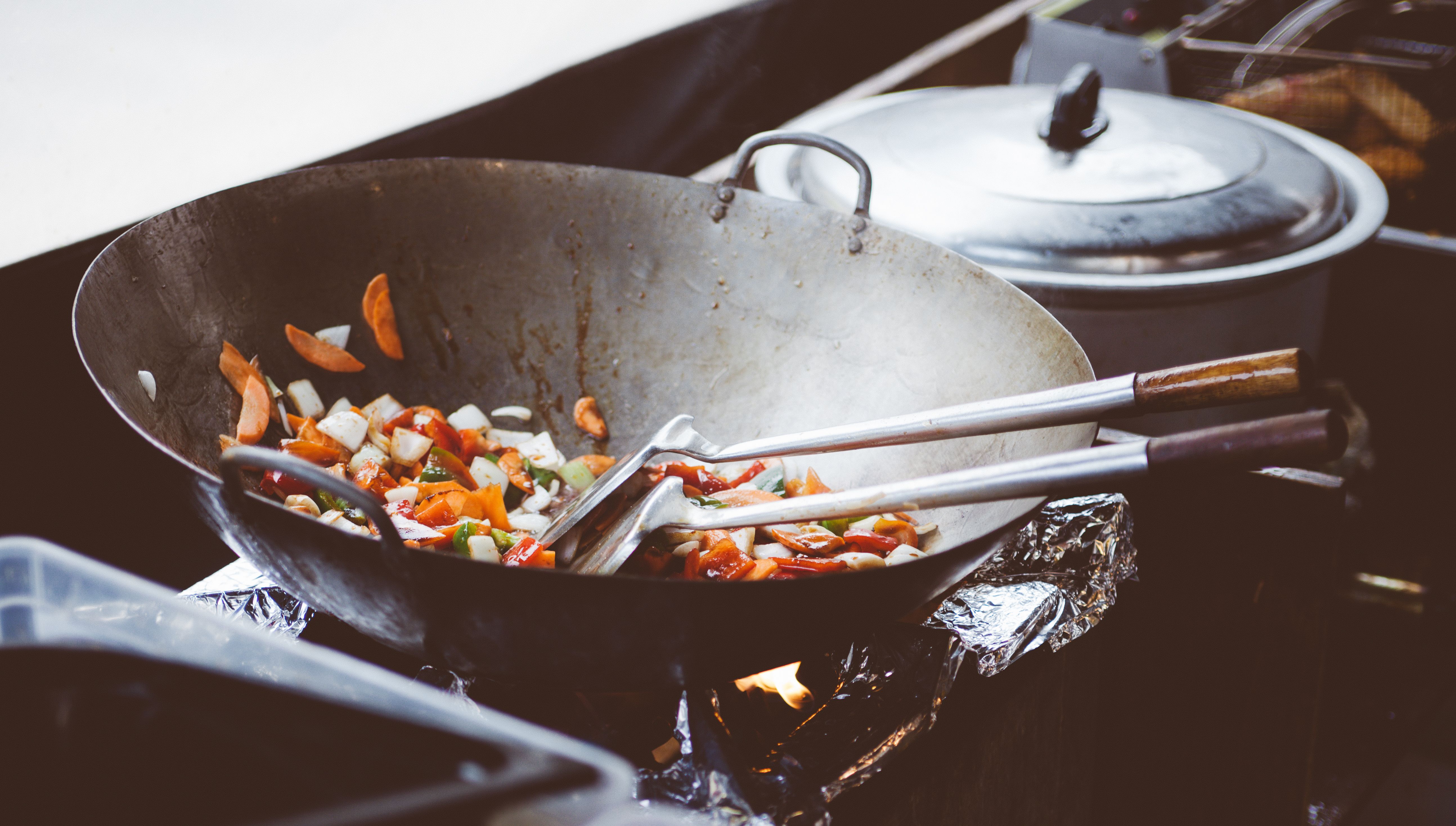 Vegetables stir-frying over an open flame in a large metal wok with cooking utensils resting inside.