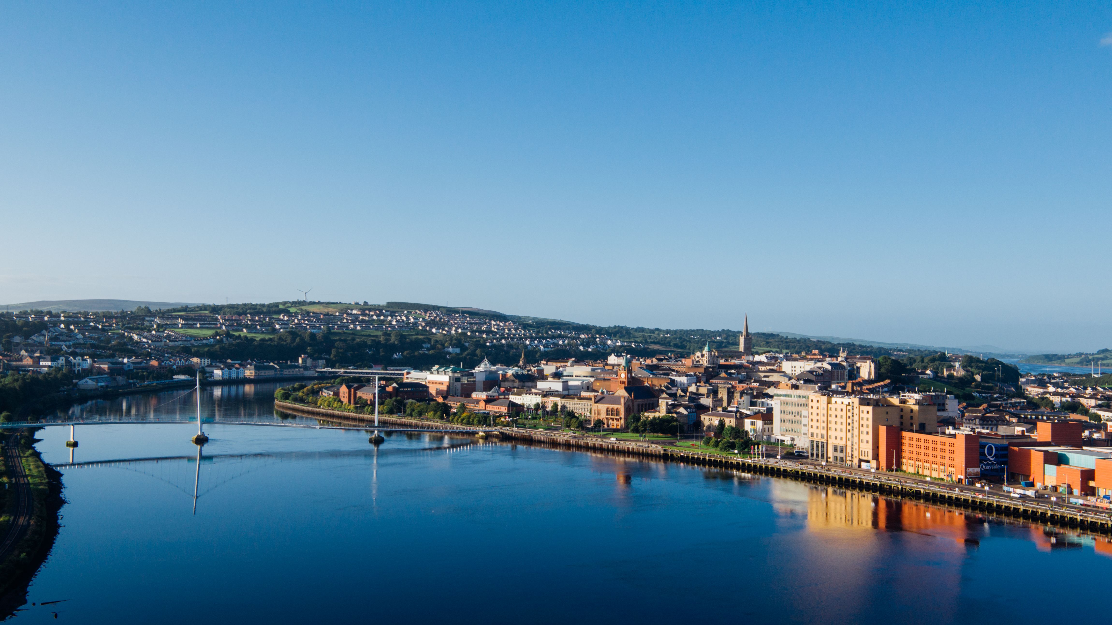 Panoramic high-angle view of a city waterfront skyline featuring a wide river crossed by a pedestrian bridge under a clear blue sky.
