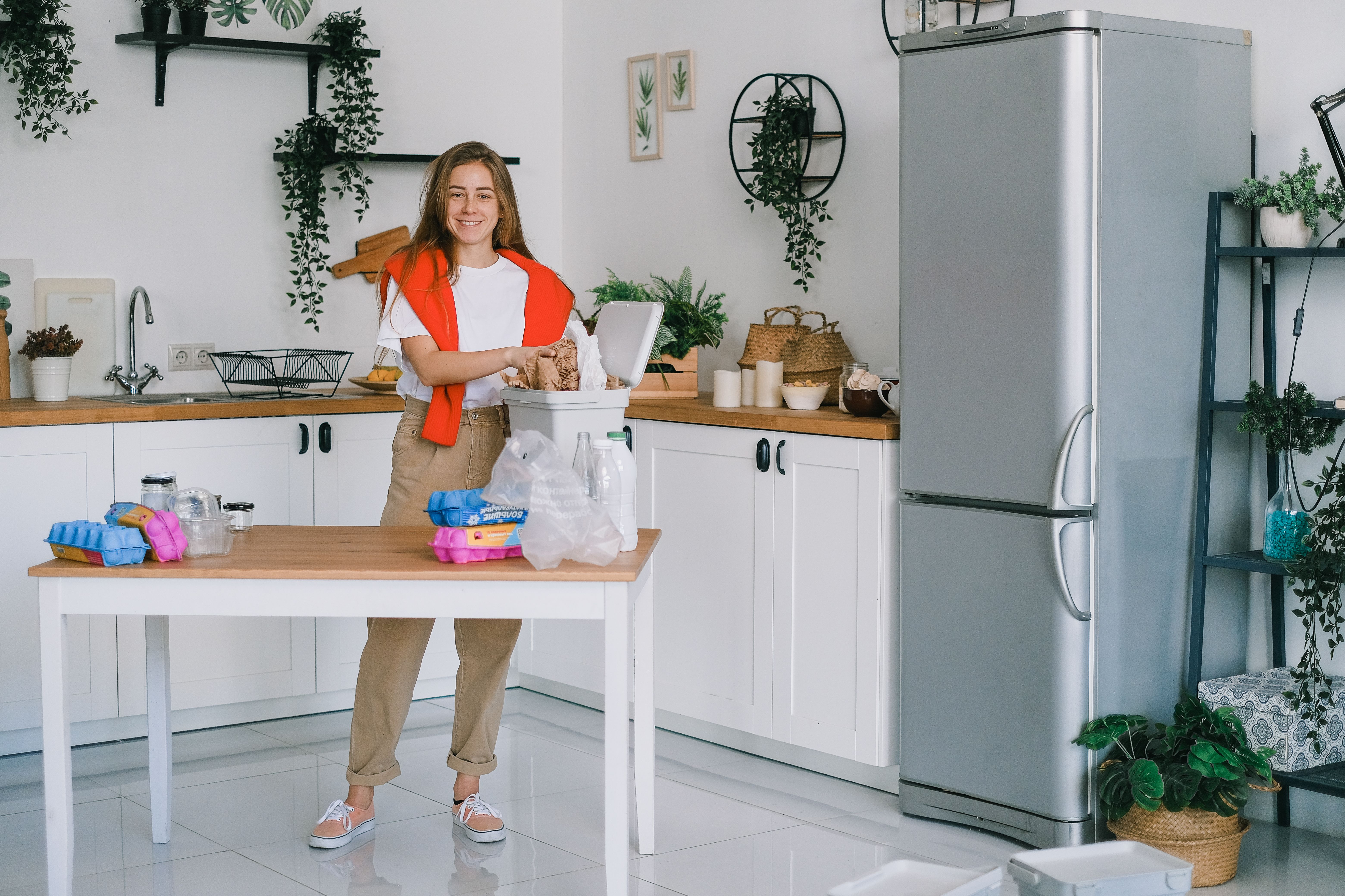 A smiling woman in a white t-shirt and red sweater sorting household waste and recyclable packaging into a small bin in a modern white kitchen.