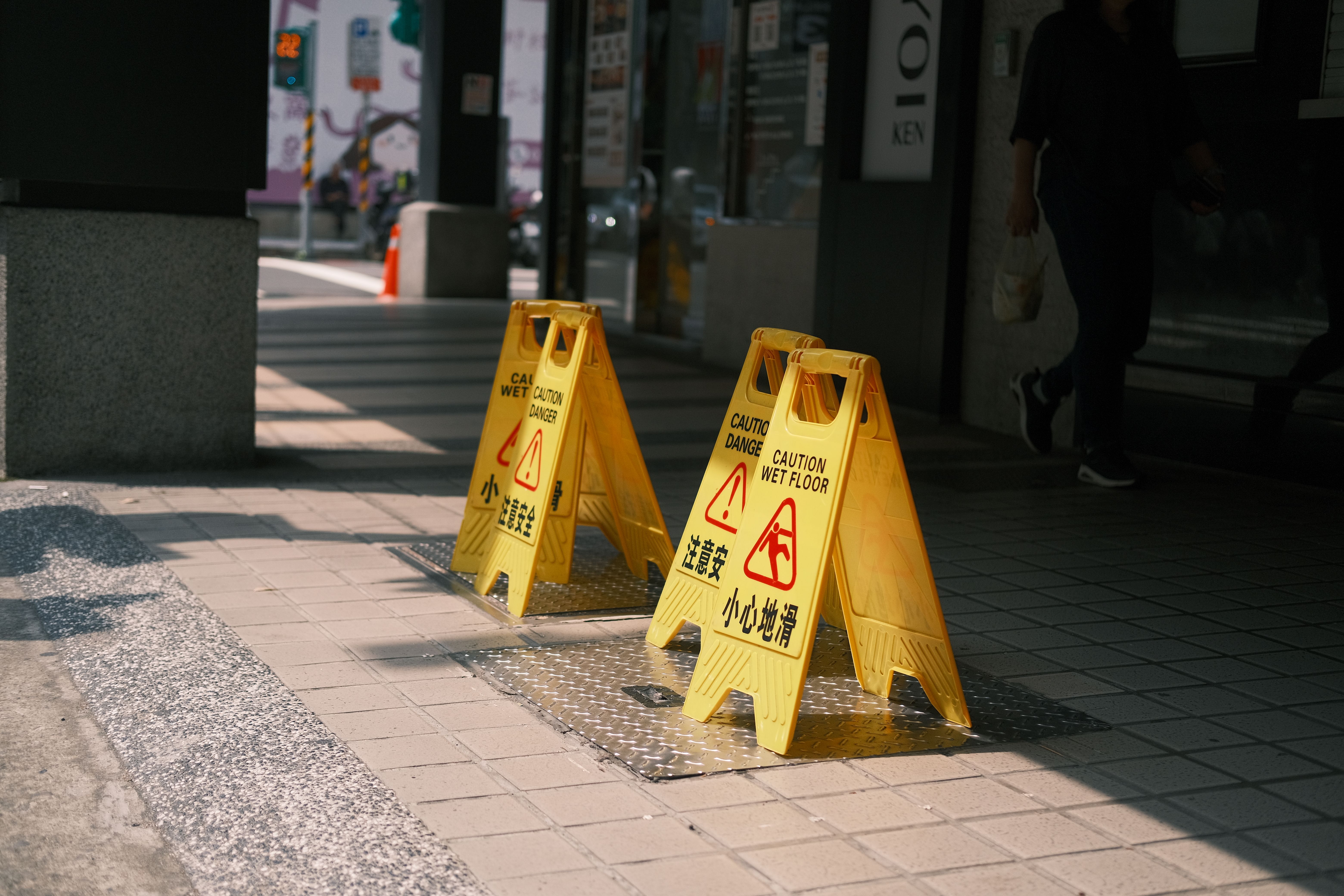 Bright yellow "Caution Wet Floor" warning signs positioned on a tiled sidewalk outside a commercial entrance.