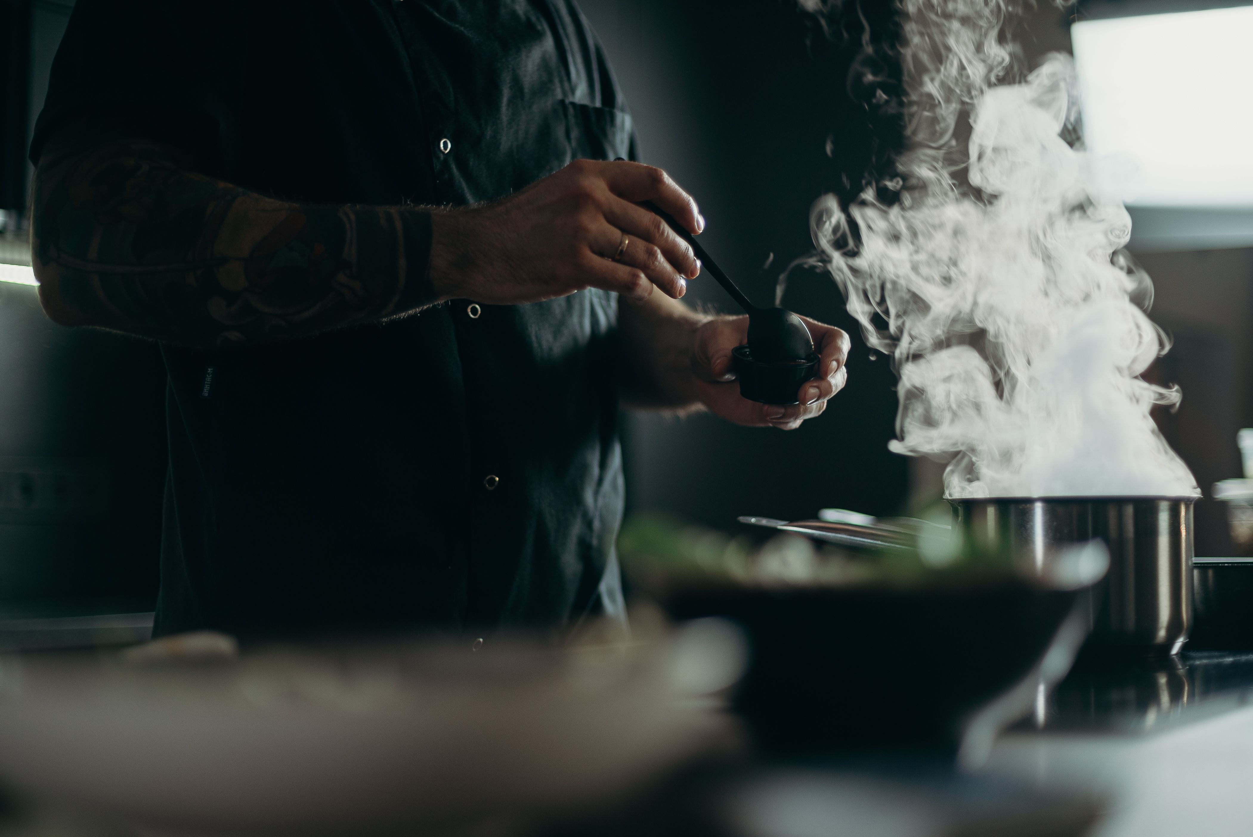 Close-up of a chef with a tattooed arm ladling liquid from a steaming pot on a stove in a kitchen.
