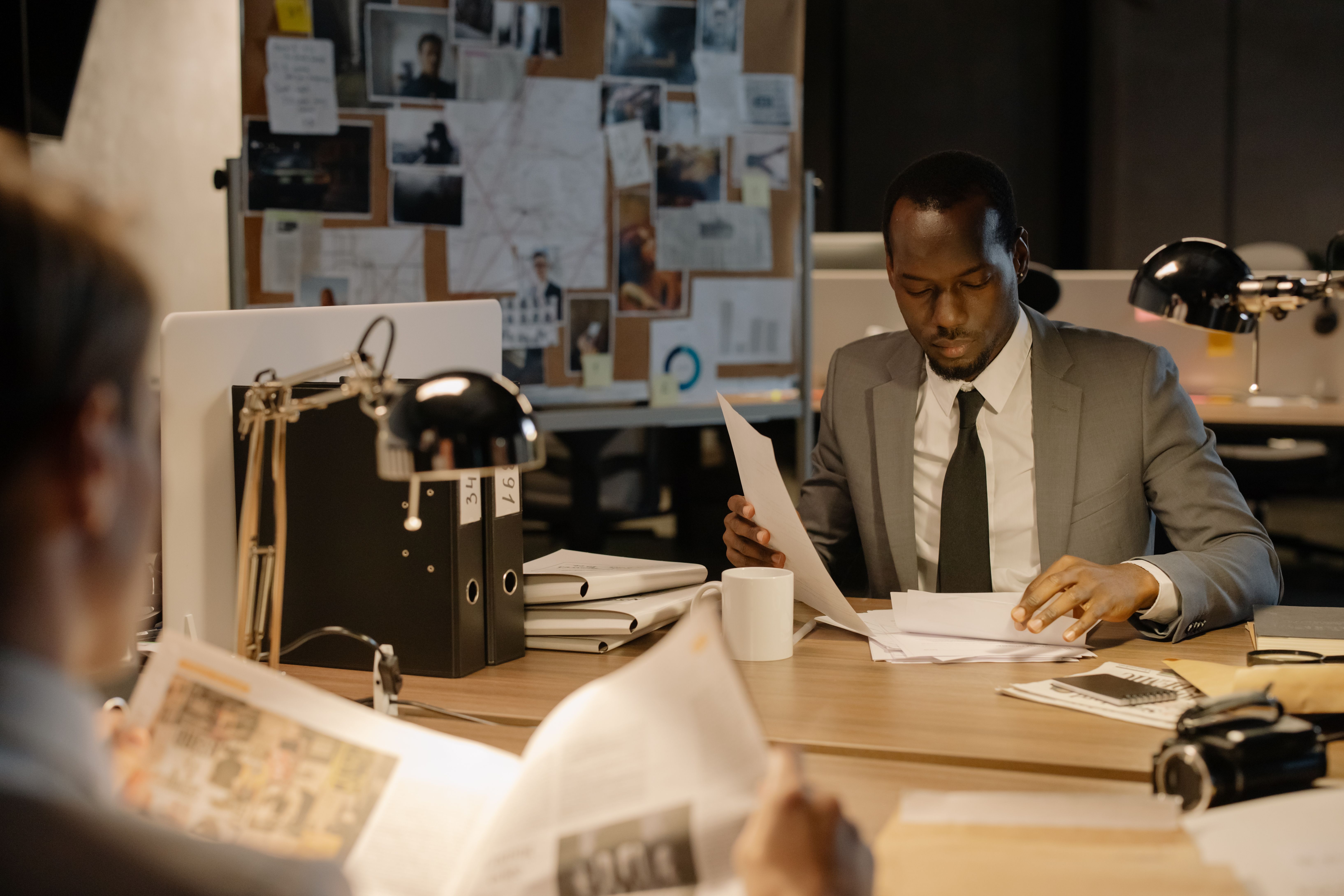 A man in a suit seated at an office desk carefully reviewing paperwork beside a desk lamp and stacked files.