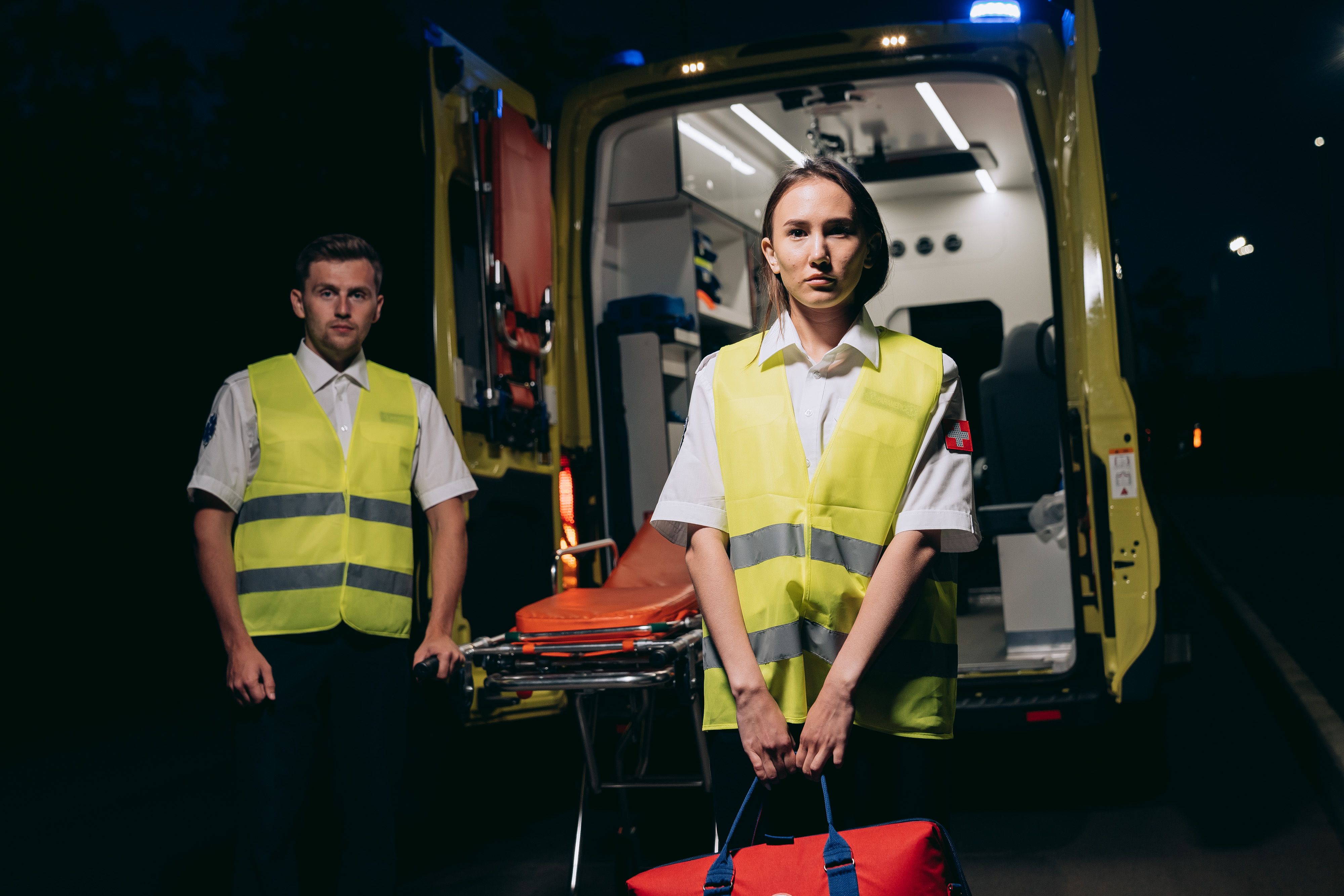 Two paramedics in high-visibility vests standing in front of an open ambulance with a stretcher visible inside.