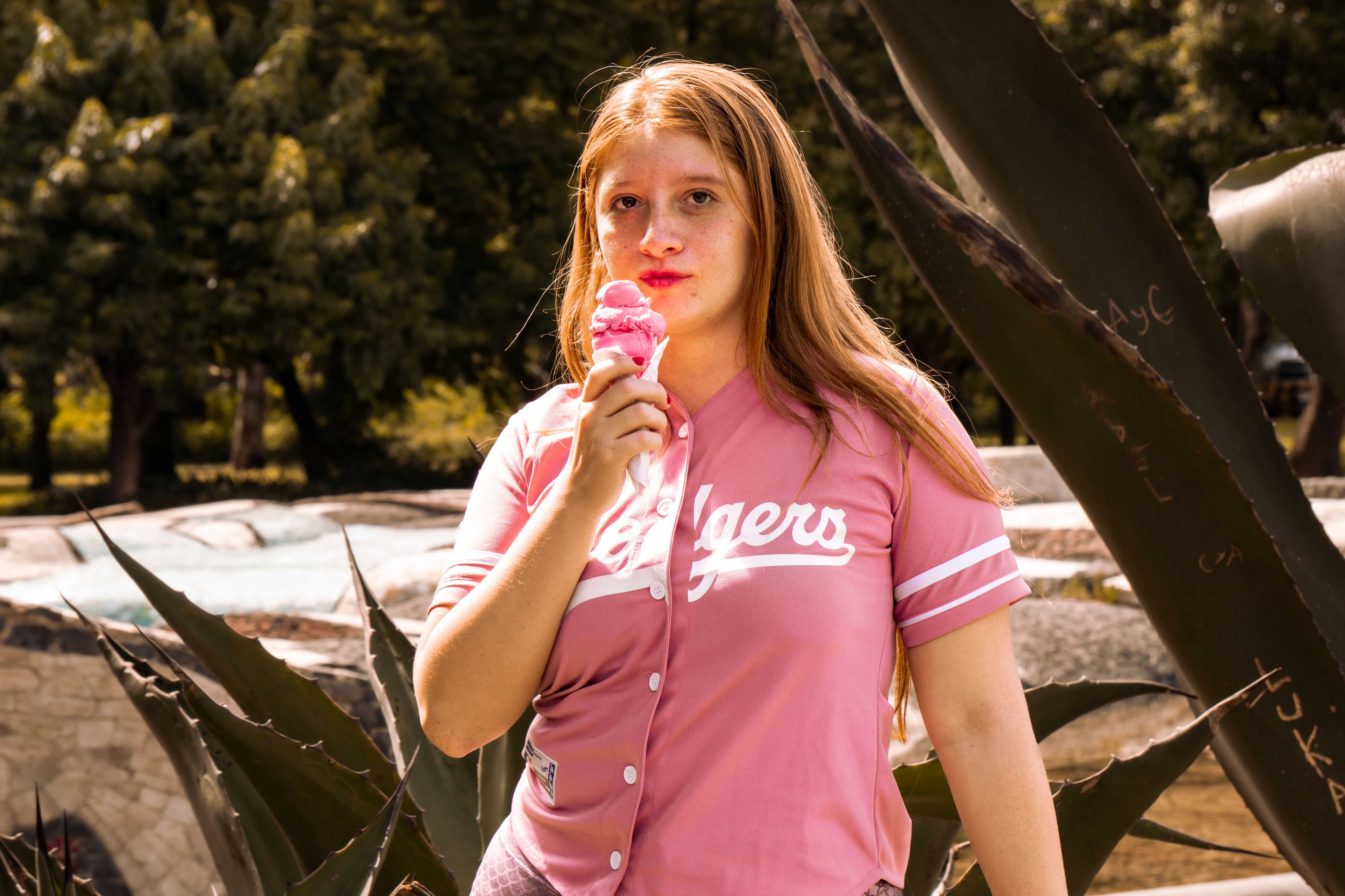 A young woman wearing a pink baseball jersey eating a pink ice cream cone outdoors near large agave plants.